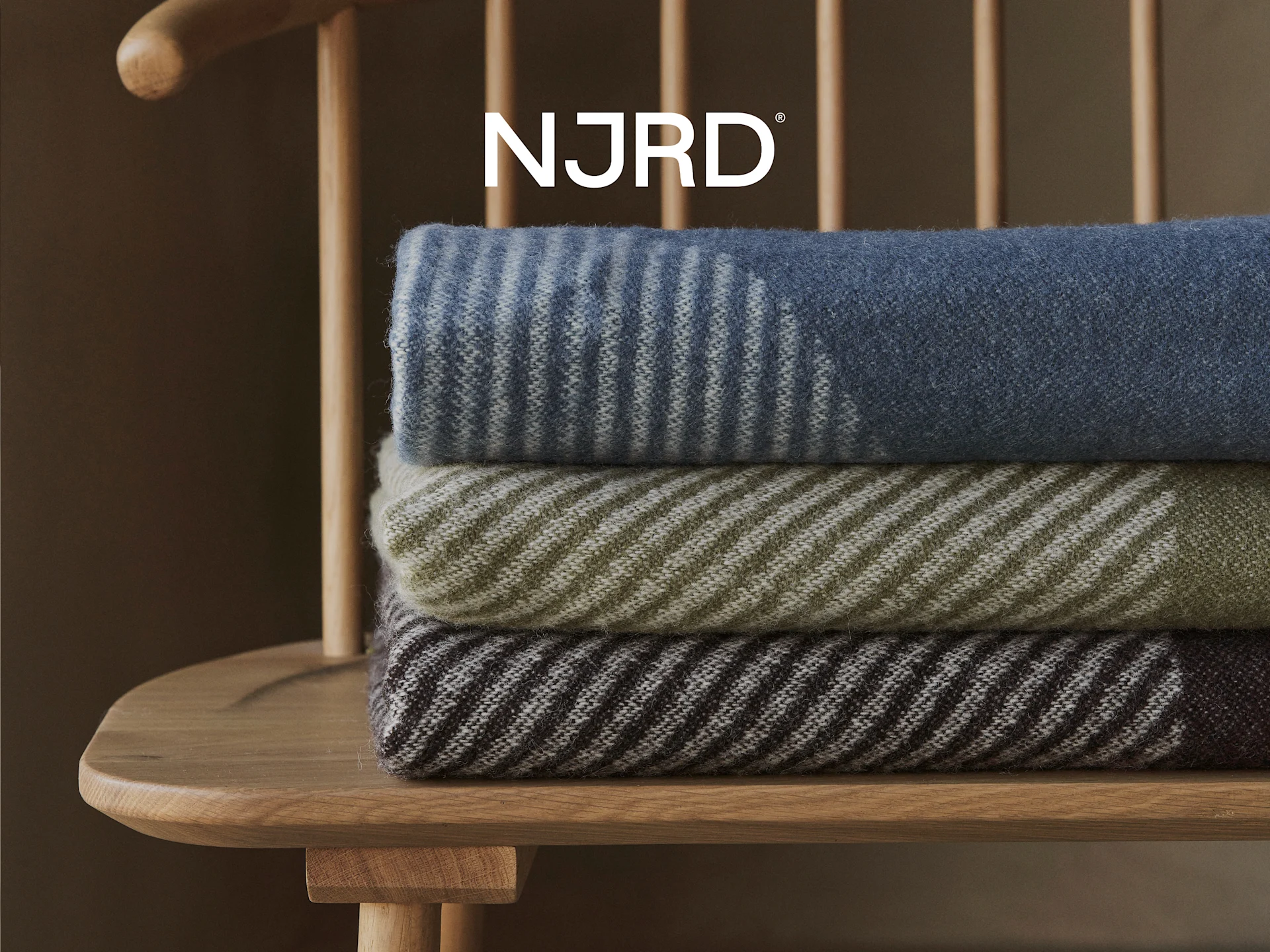 Tre stripete tepper i blått, oliven og brunt, stablet på en trestol med NJRD-logoen over.