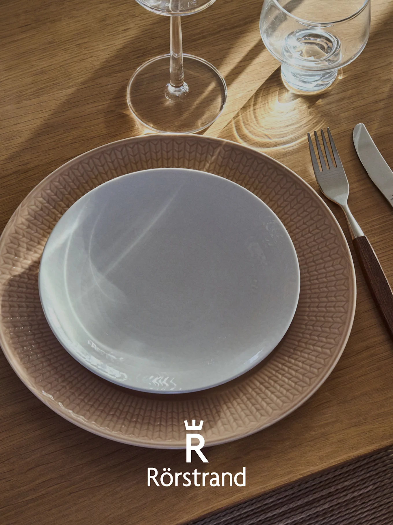En borddekking med en hvit tallerken på en mønstret beige tallerken, en gaffel, en kniv og to glass på et trebord. Rörstrand-logo.