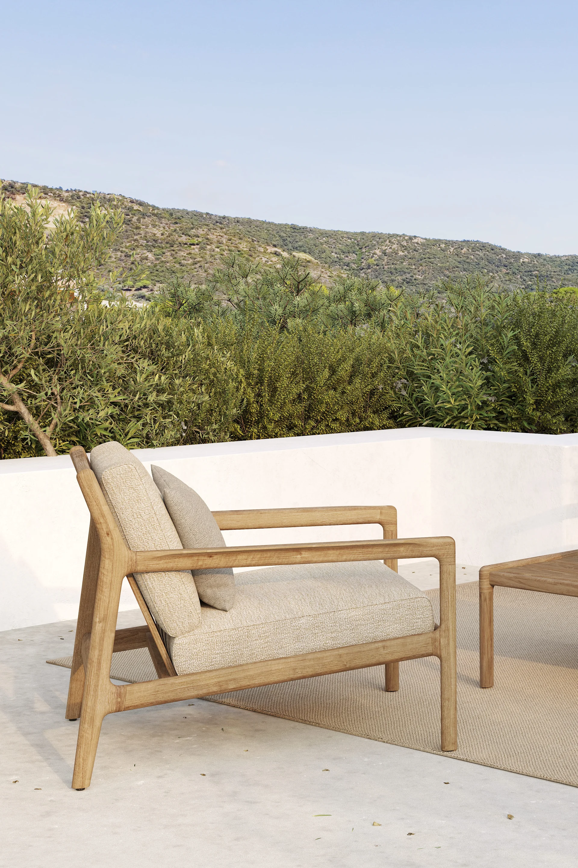 Utendørs lenestol i tre med lyse puter på en uteplass, mot grønne åser og en blå himmel.