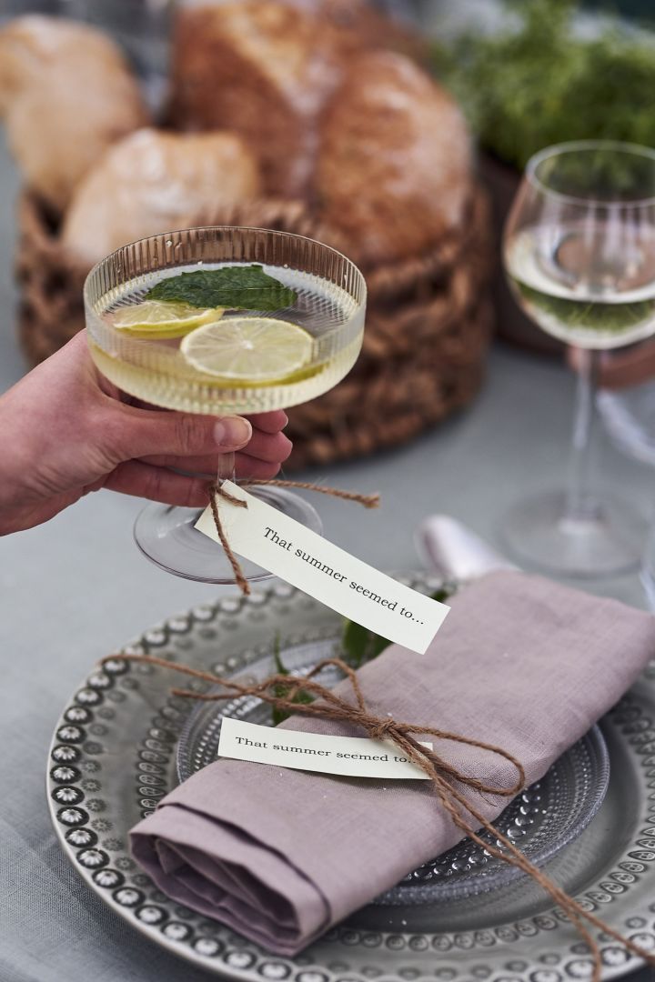 Sommerlig bordkort i form av en sommersang på lapp knytt rundt champagneglasset og servietten på bordet.