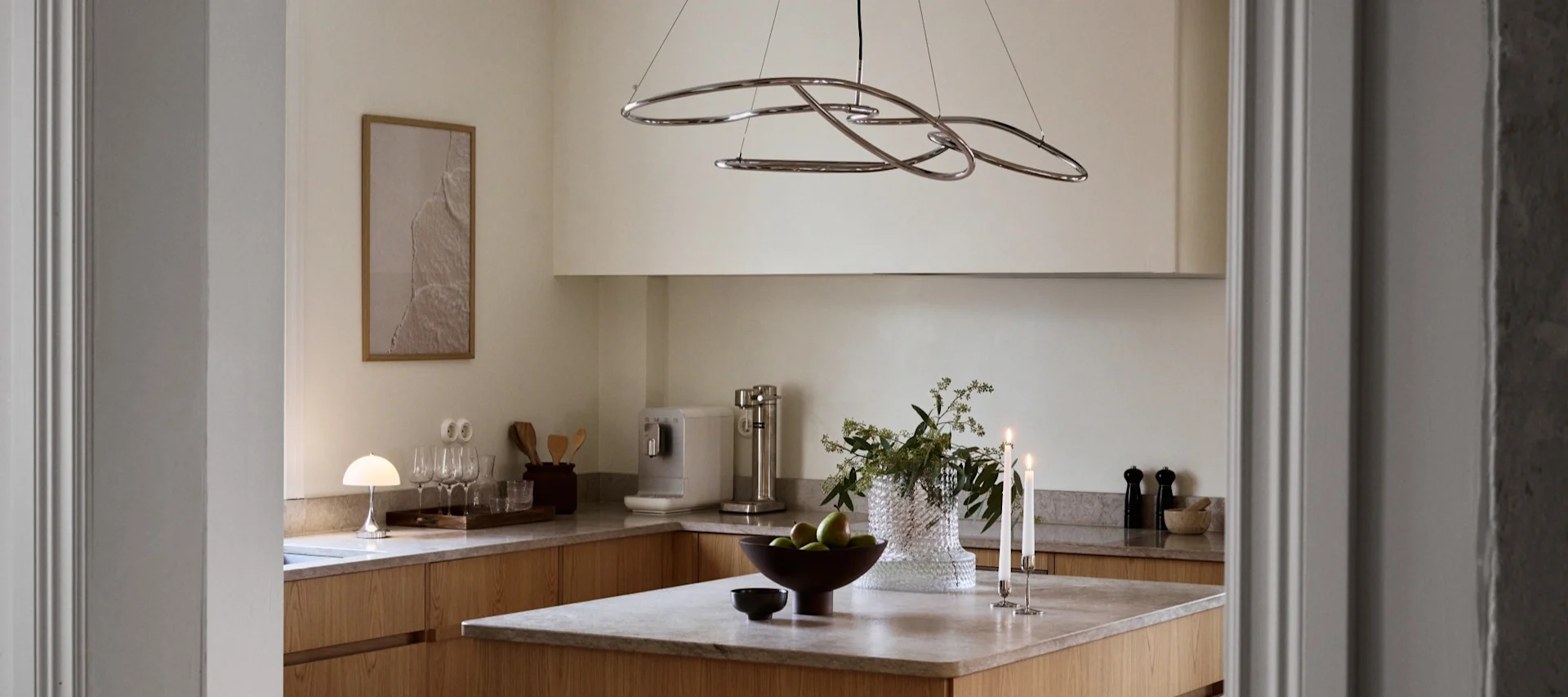 Moderne kjøkken med lyse treverksskap, øy med epler, tente lys og en skulpturell kromlysekrone.