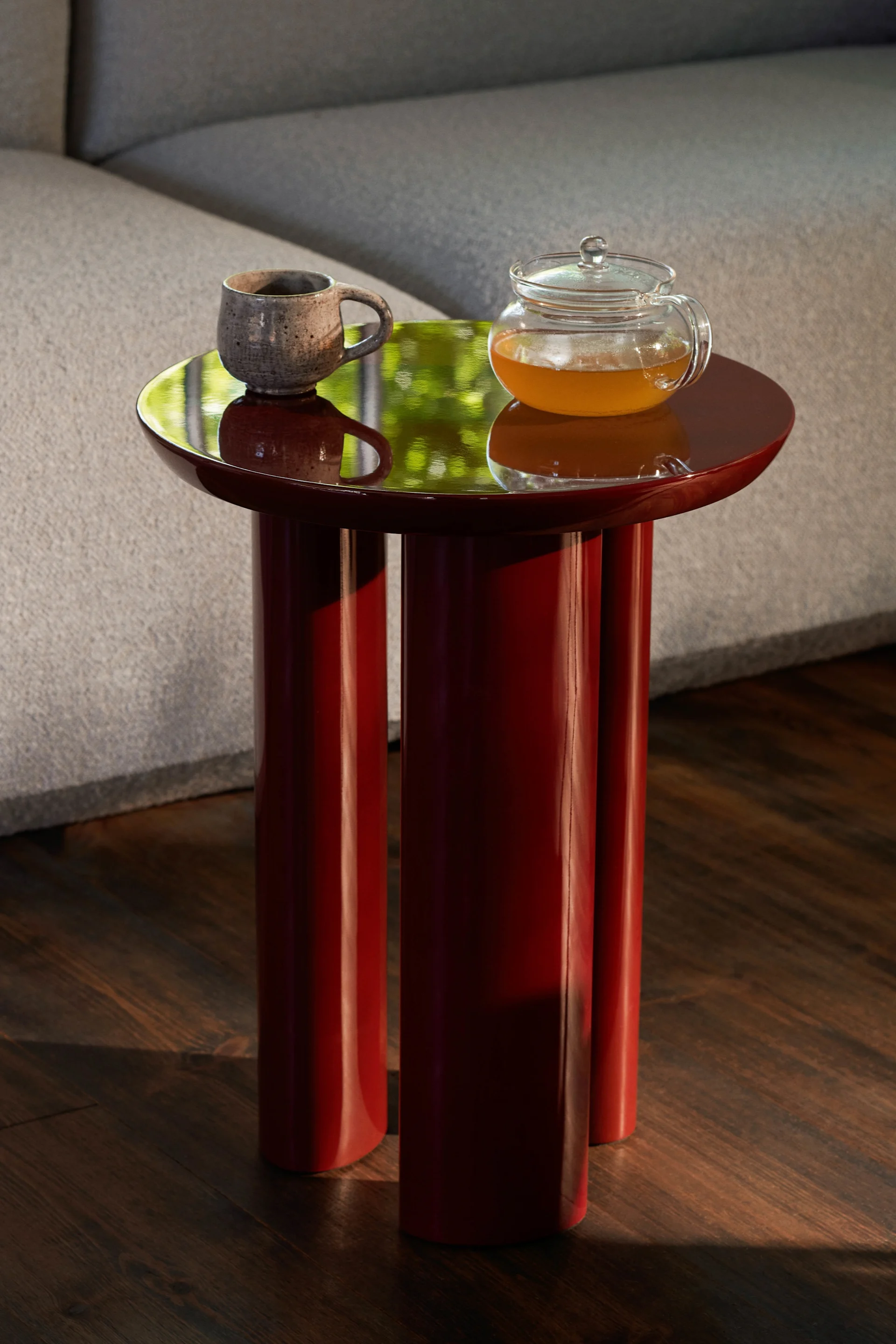 Mørkerødt glansfullt sidebord med krus og tekanne. Grønne refleksjoner på toppen, ved en grå sofa og tregulv.