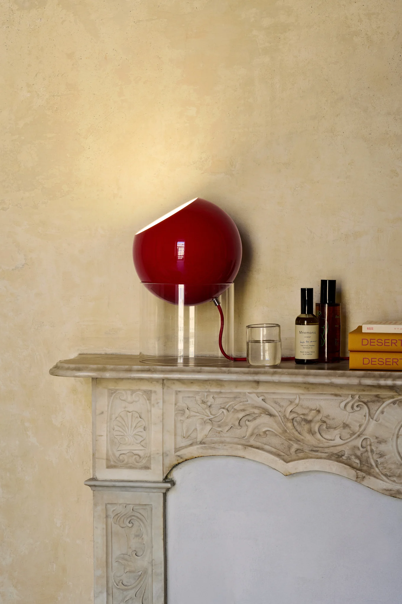 Rød sfærisk lampe med gjennomsiktig base på dekorativ marmorkaminhylle, ved siden av et glass vann, to flasker og to gule bøker.