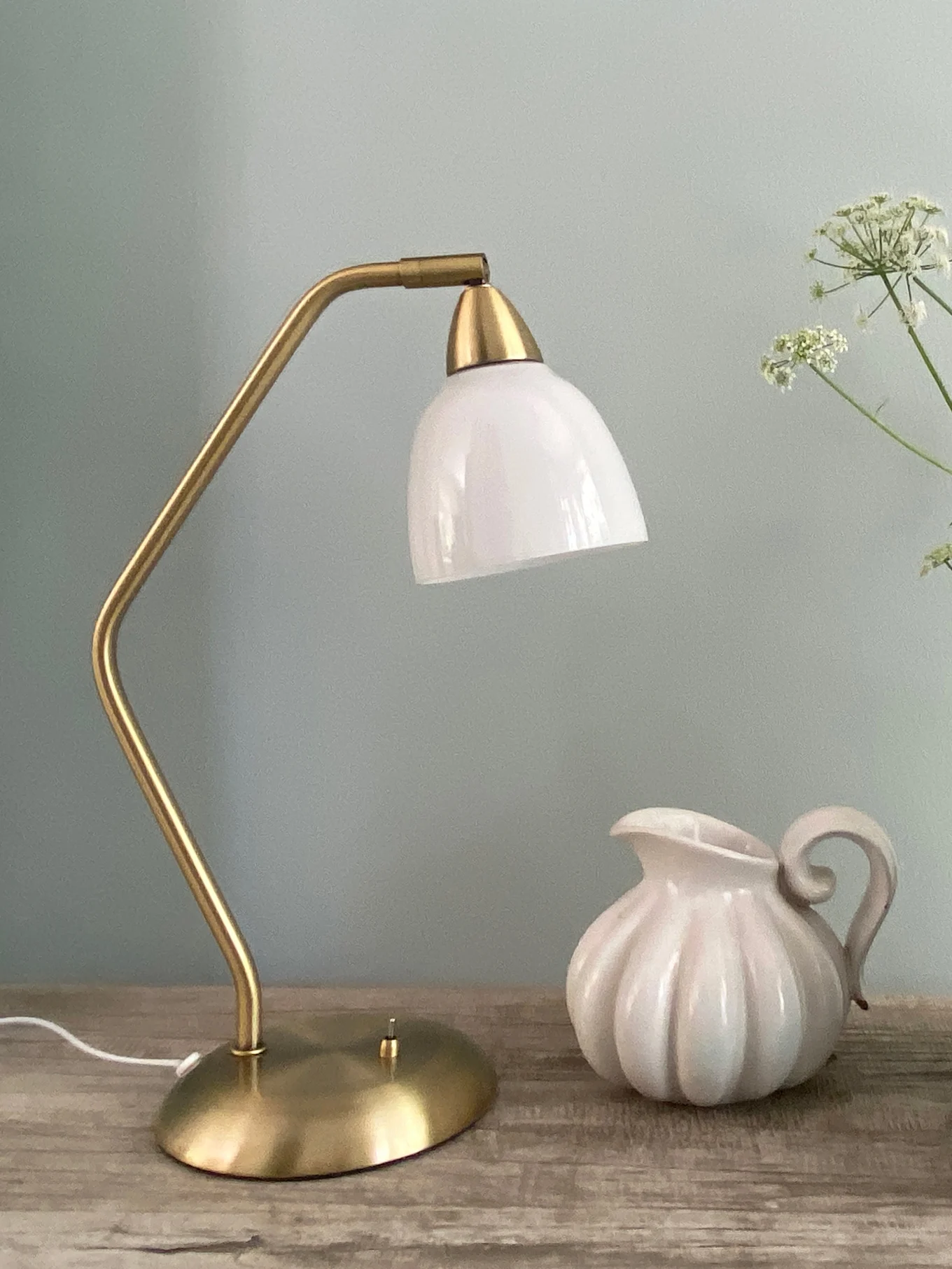 En messing bordlampe med en hvit glasskjerm står ved siden av en hvit riflet keramisk kanne på en treoverflate.