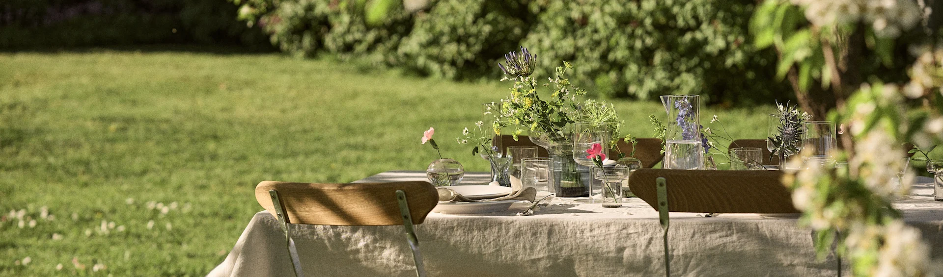 Bord dekket for et utendørs måltid i en hage, med duk, servise, glass og blomsteroppsatser.