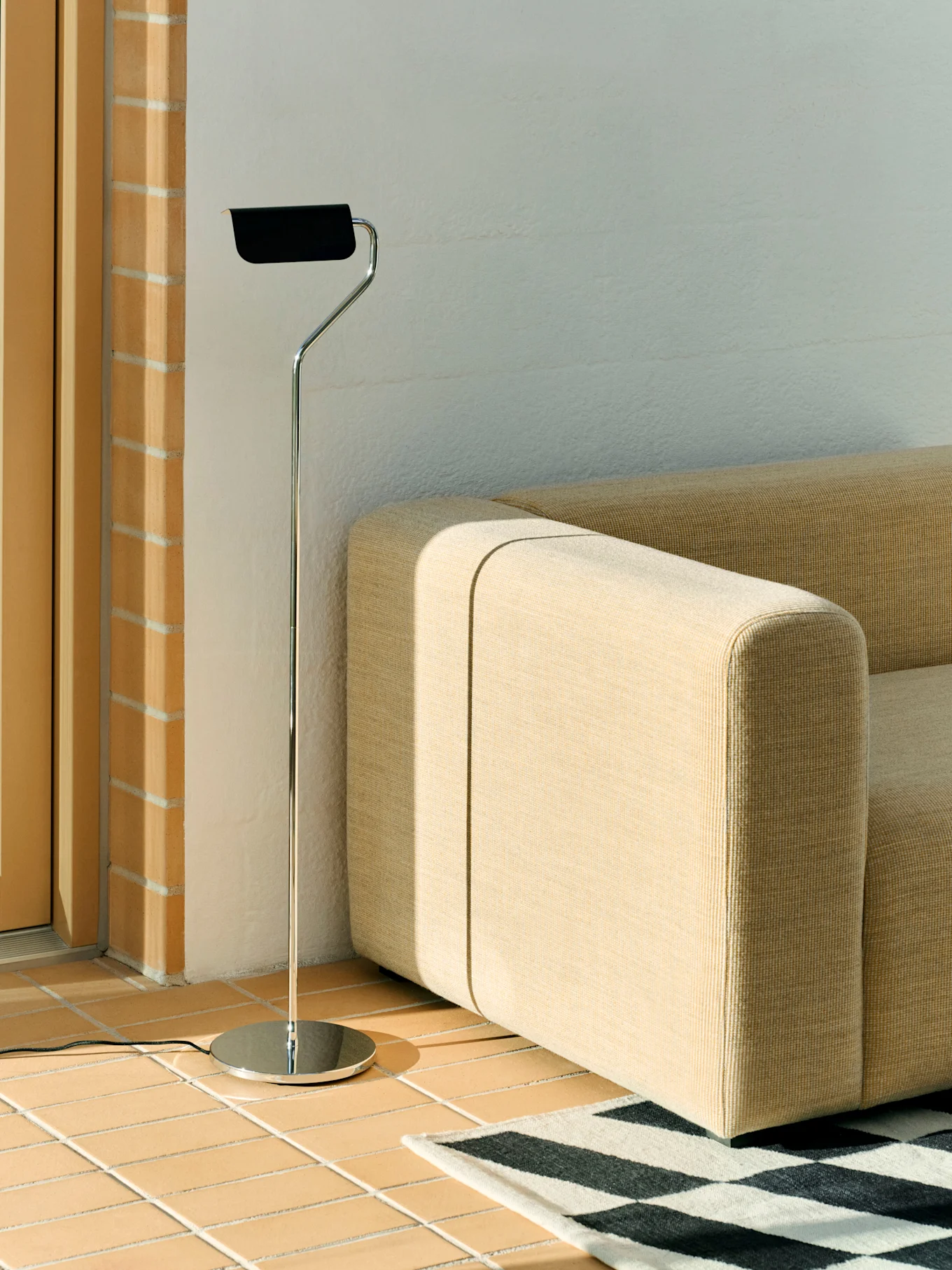 En gulvlampe i krom med svart skjerm står ved siden av en beige modulsofa og et svart-hvitt geometrisk teppe på et flislagt gulv.
