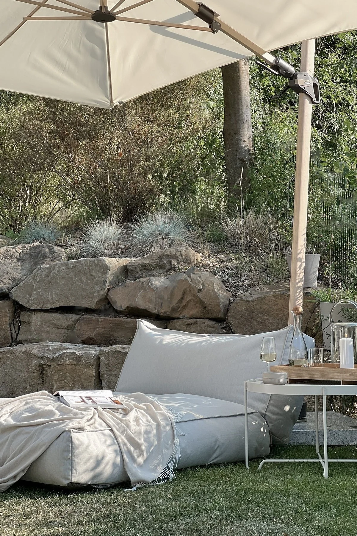 Avslappet uteområde med stor parasoll, to lyse gulvputer, et pledd, et magasin og et sidebord med drikkevarer.