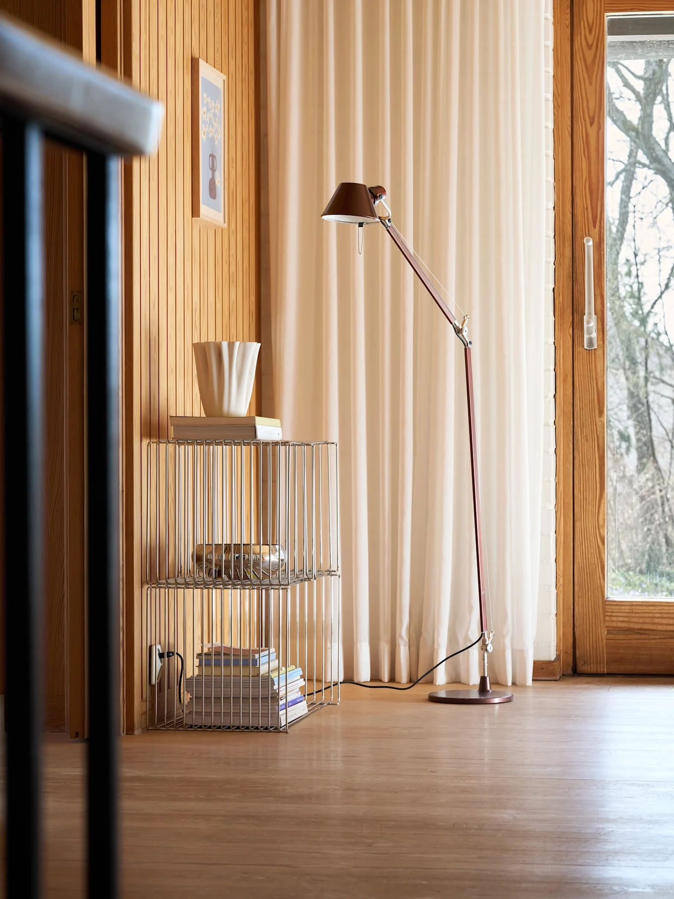 Et koselig rom med trevegger og -gulv, en rød gulvlampe, en kromhylle med bøker og en riflet hvit vase, samt et vindu med lyse gardiner.
