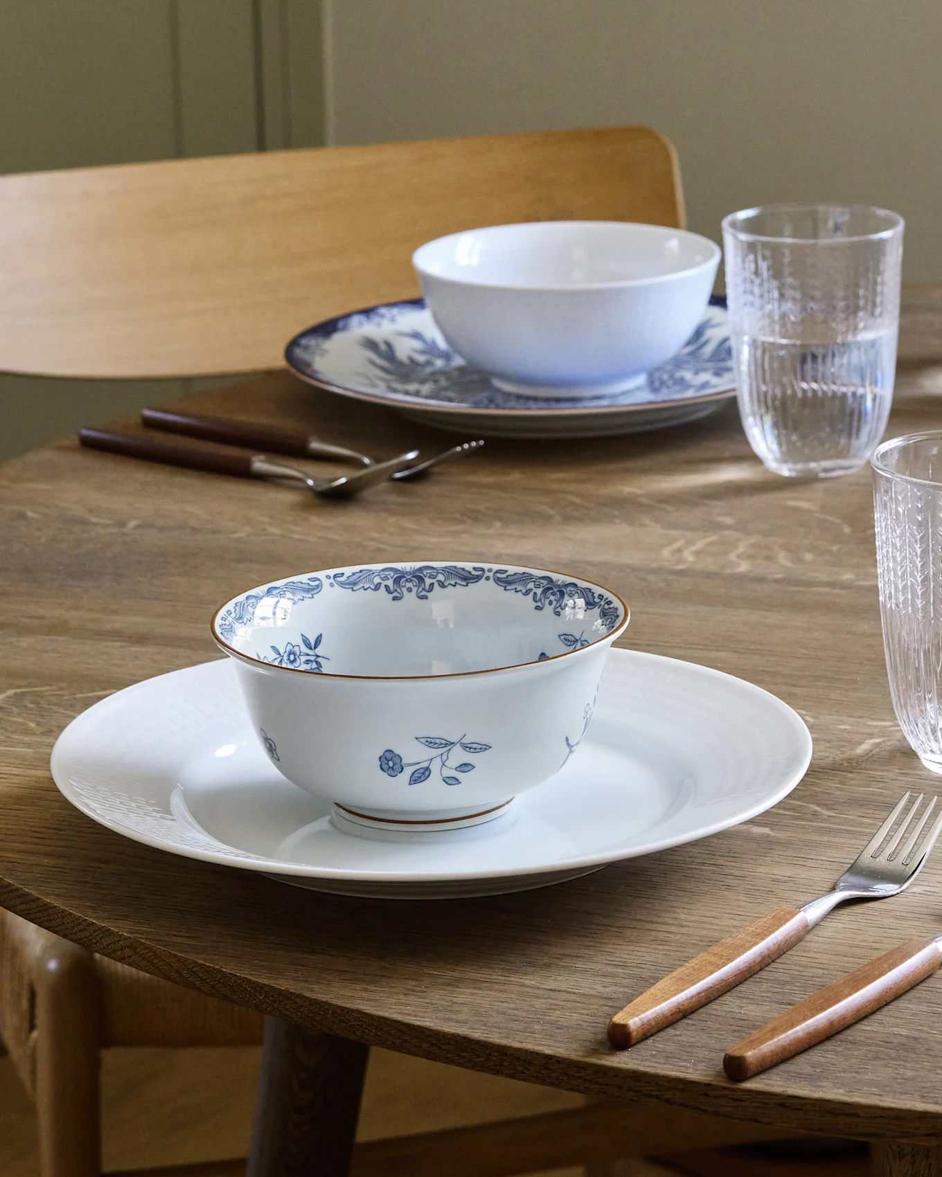 Spisebord dekket for to med hvite og blåmønstrede skåler på hvite tallerkener, bestikk med trehåndtak og vannglass.