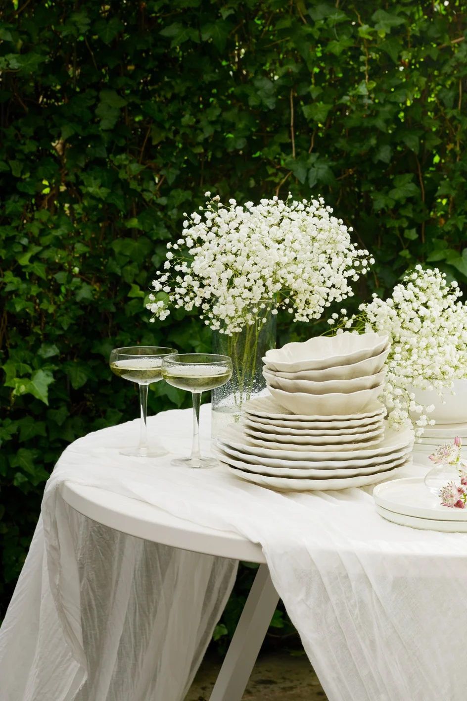 Utendørs bord dekket med hvitt servise, champagneglass og brudeslørblomster mot grønt løv.