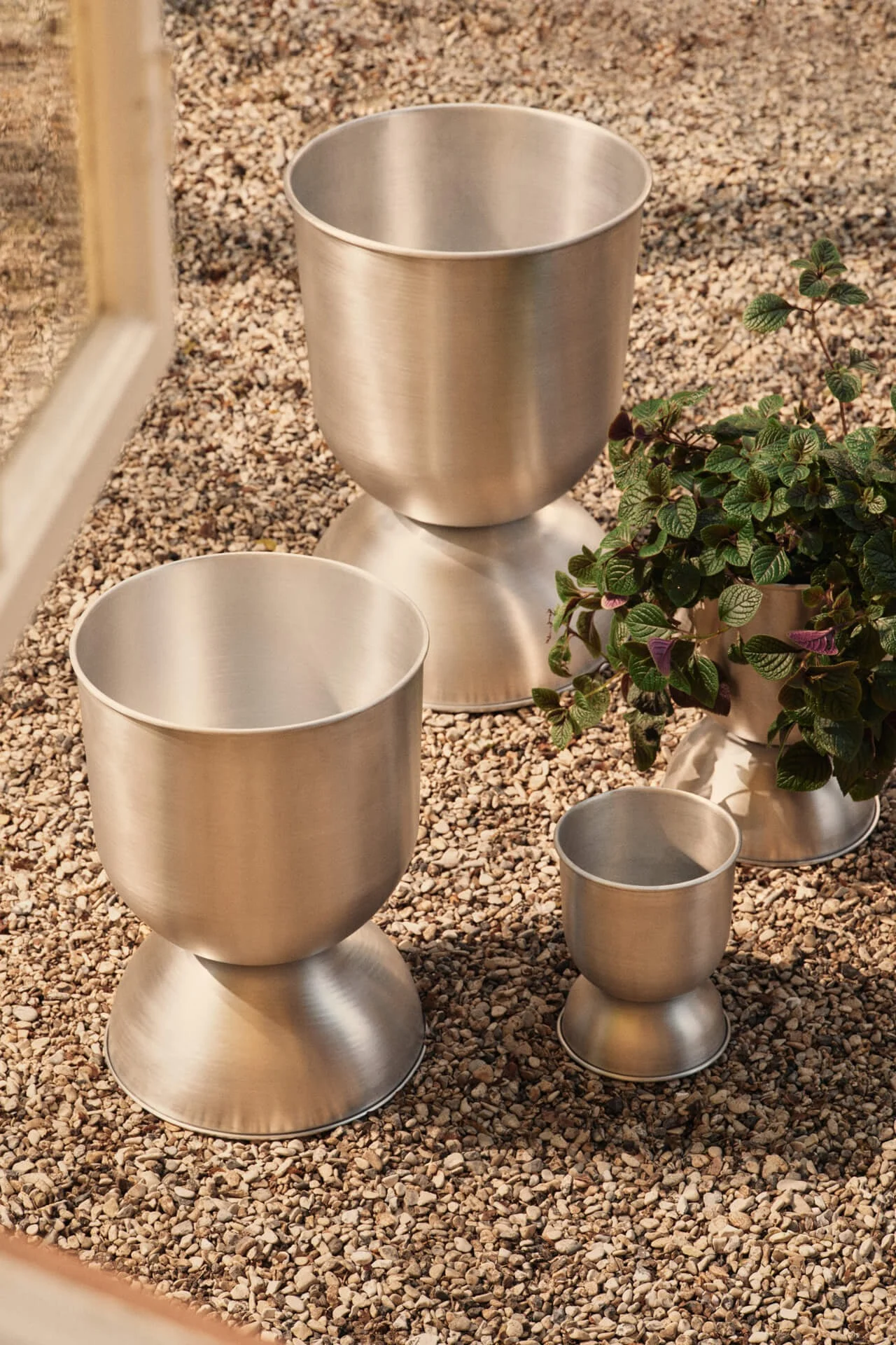 Tre børstede metallplantere i forskjellige størrelser på småstein, en med en grønn plante.