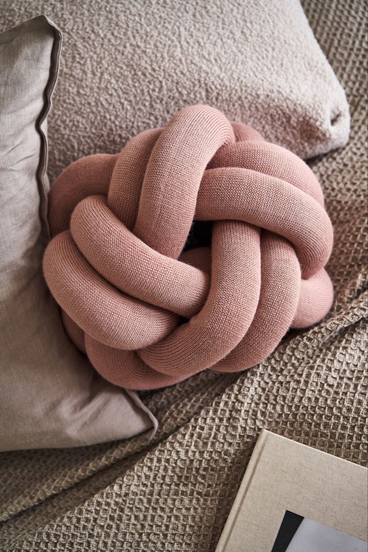 Knot pute liten i dusty pink sitter på en seng med beige og grå laken. 