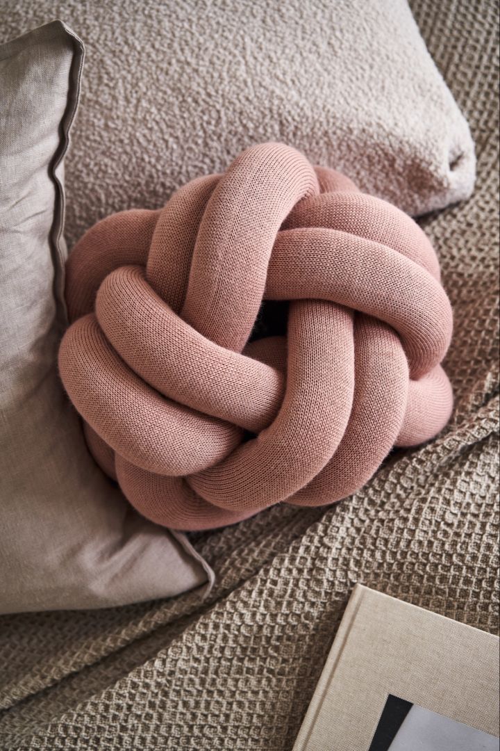 Knot pute liten i dusty pink sitter på en seng med beige og grå laken. 
