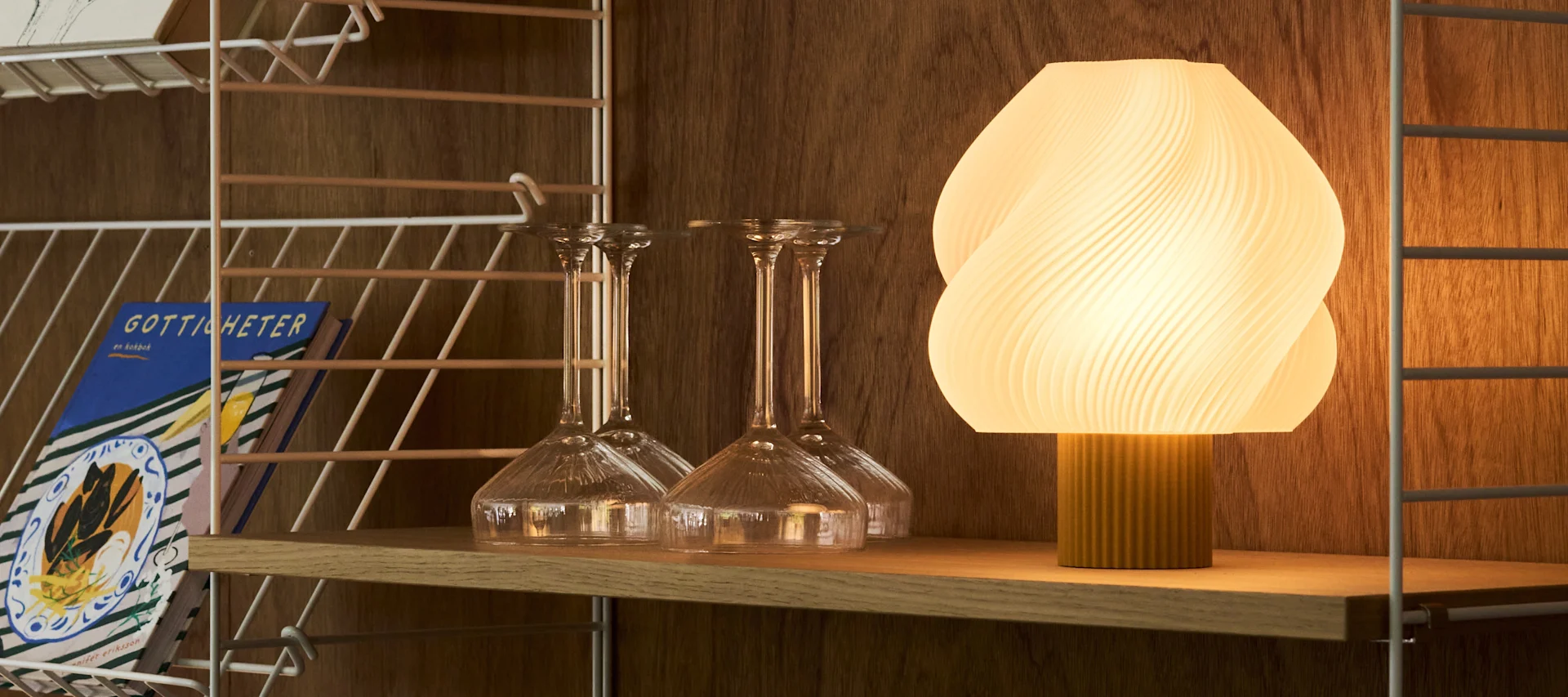 En opplyst, ribbet oval lampe og ulike klare glass står på en trebarvogn mot en varm, teksturert vegg.