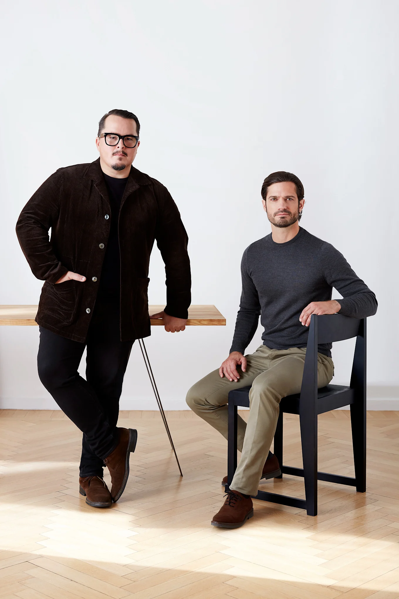 To menn, den ene står ved et trebord i en brun kordfløyelsjakke, den andre sitter på en svart stol i en grå skjorte.