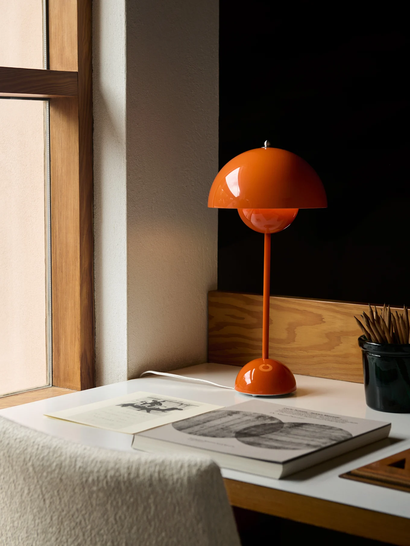 En oransje soppformet bordlampe på et hvitt skrivebord ved siden av et vindu, med bøker og blyanter.