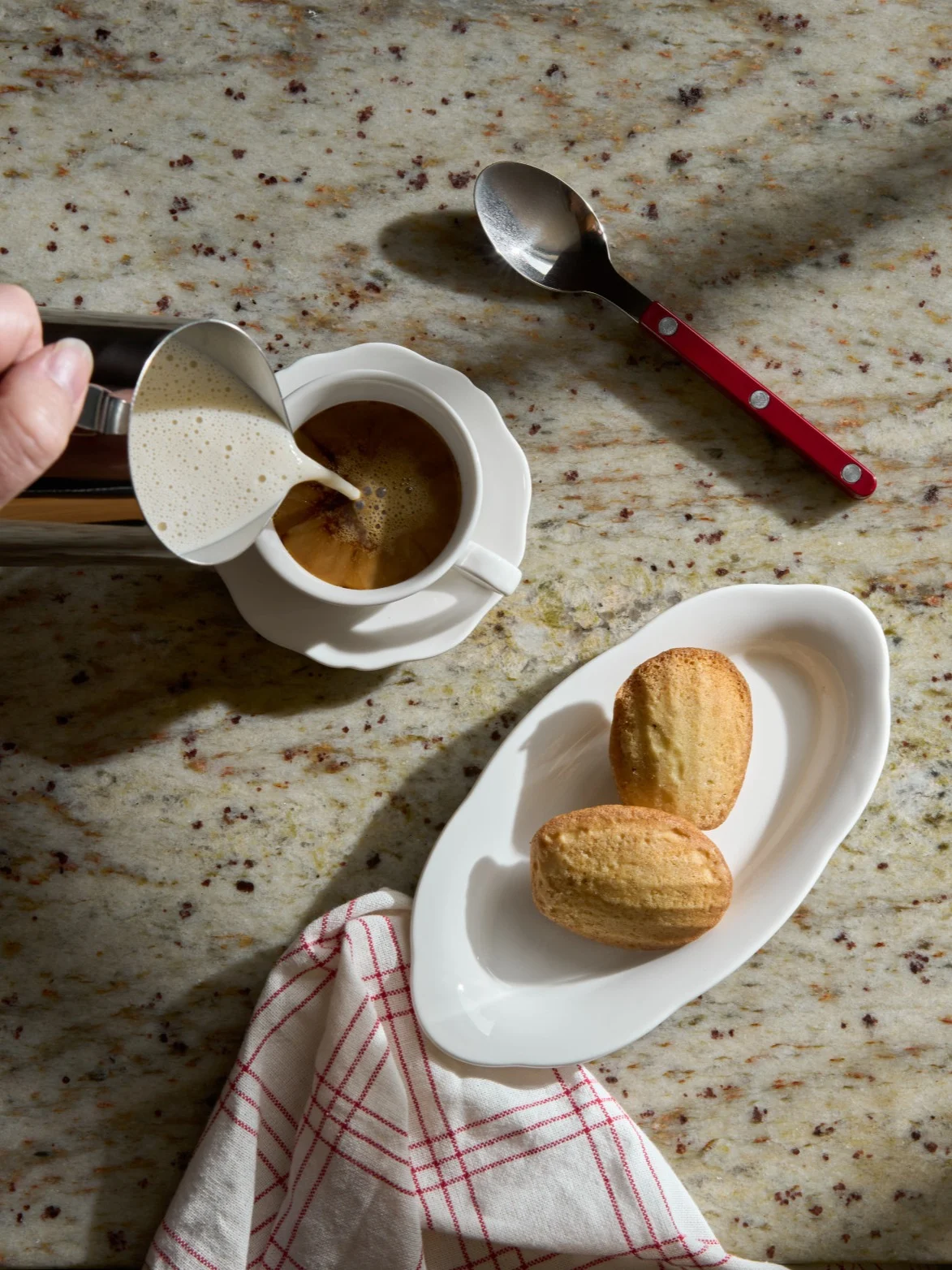 Hånd heller skummet melk i en kaffekopp ved siden av madeleinekaker på en granittbenk.