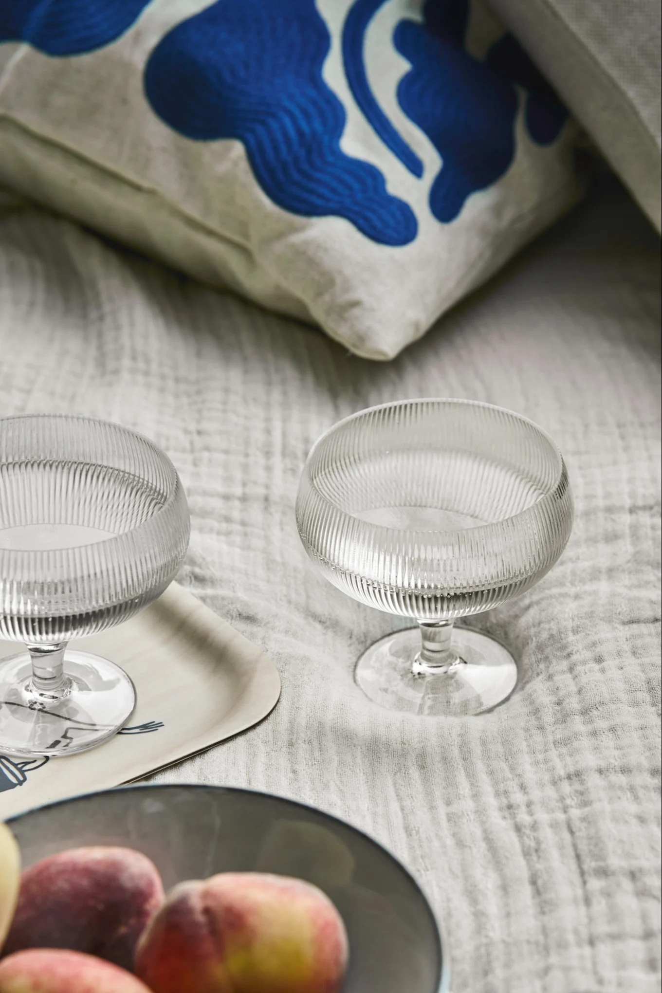 To riflede coupette-glass på et lyst brett, med fersken i en bolle og en blåmønstret pute i bakgrunnen.