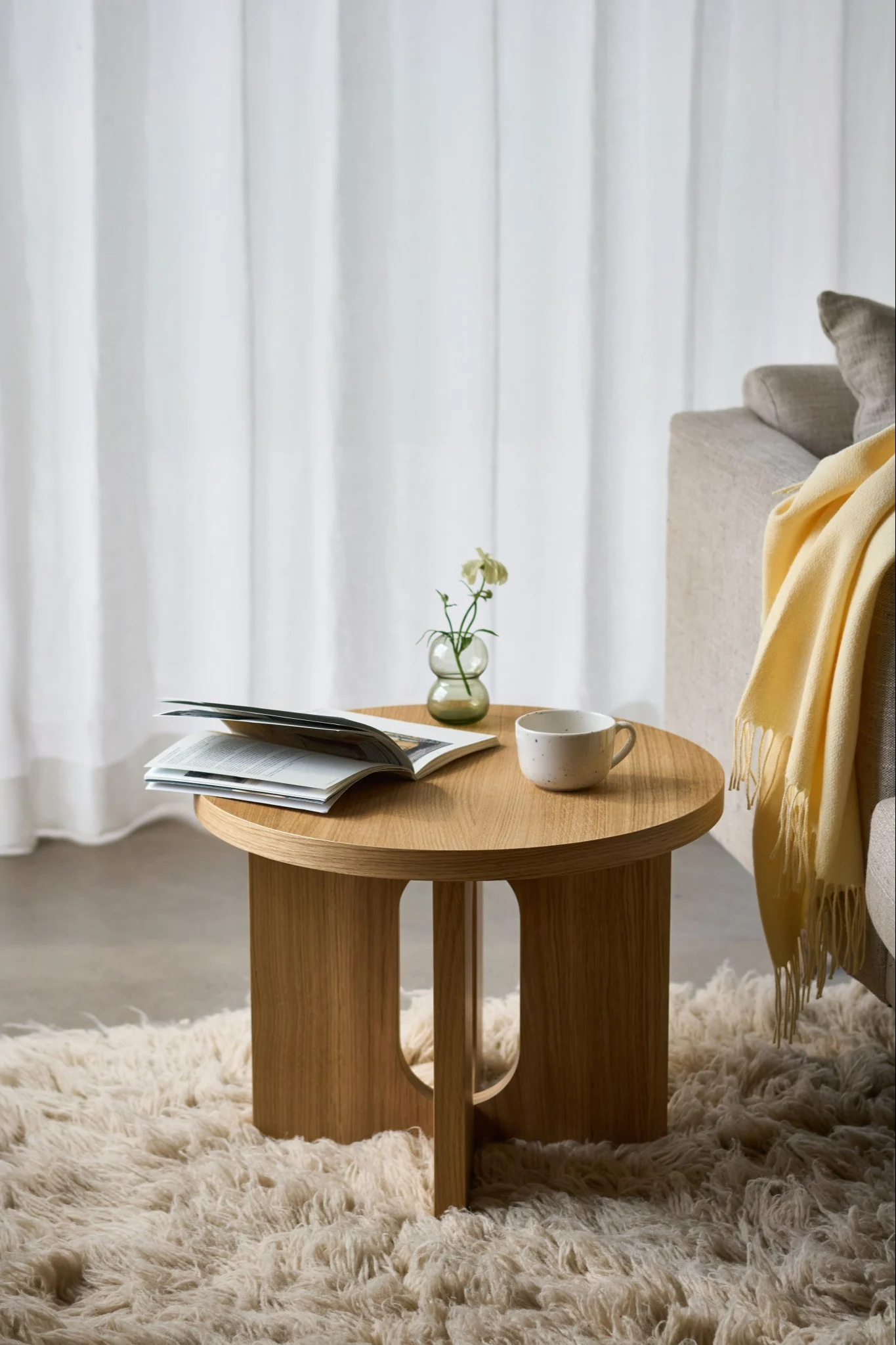 Rundt sidebord i tre med en åpen bok, et krus og en liten vase med en hvit blomst på et lodne teppe, ved siden av en sofa.