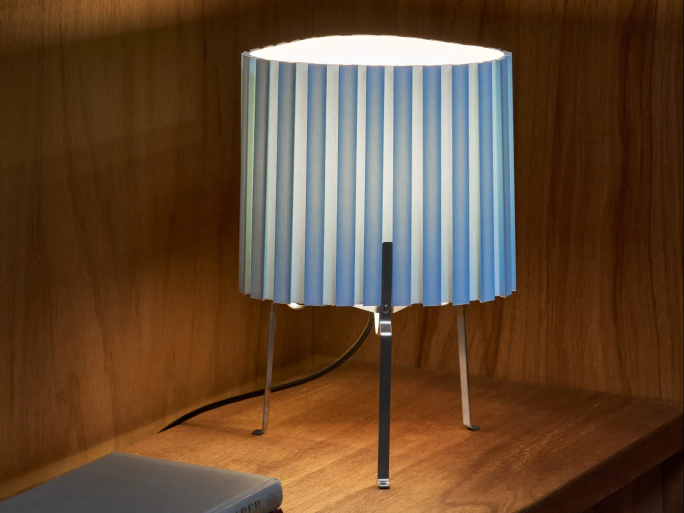 Bordlampe med plissert blå og hvit stripete skjerm på en trehylle, som lyser opp området.