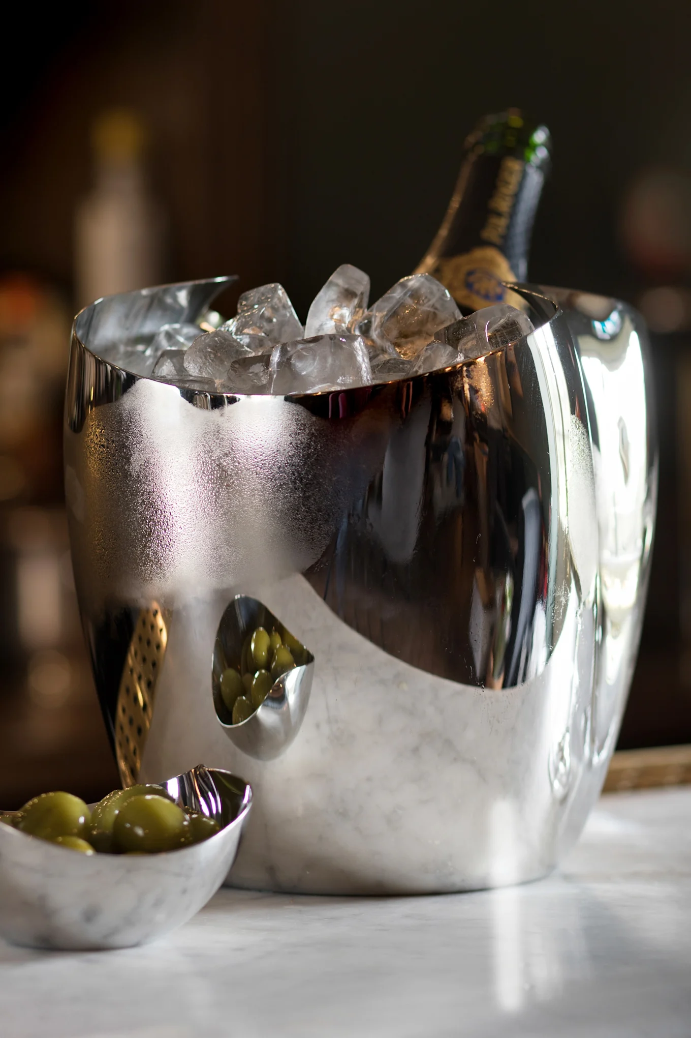 En reflekterende sølv isbøtte med en champagneflaske og is, ved siden av to små skåler med grønne oliven på marmor.