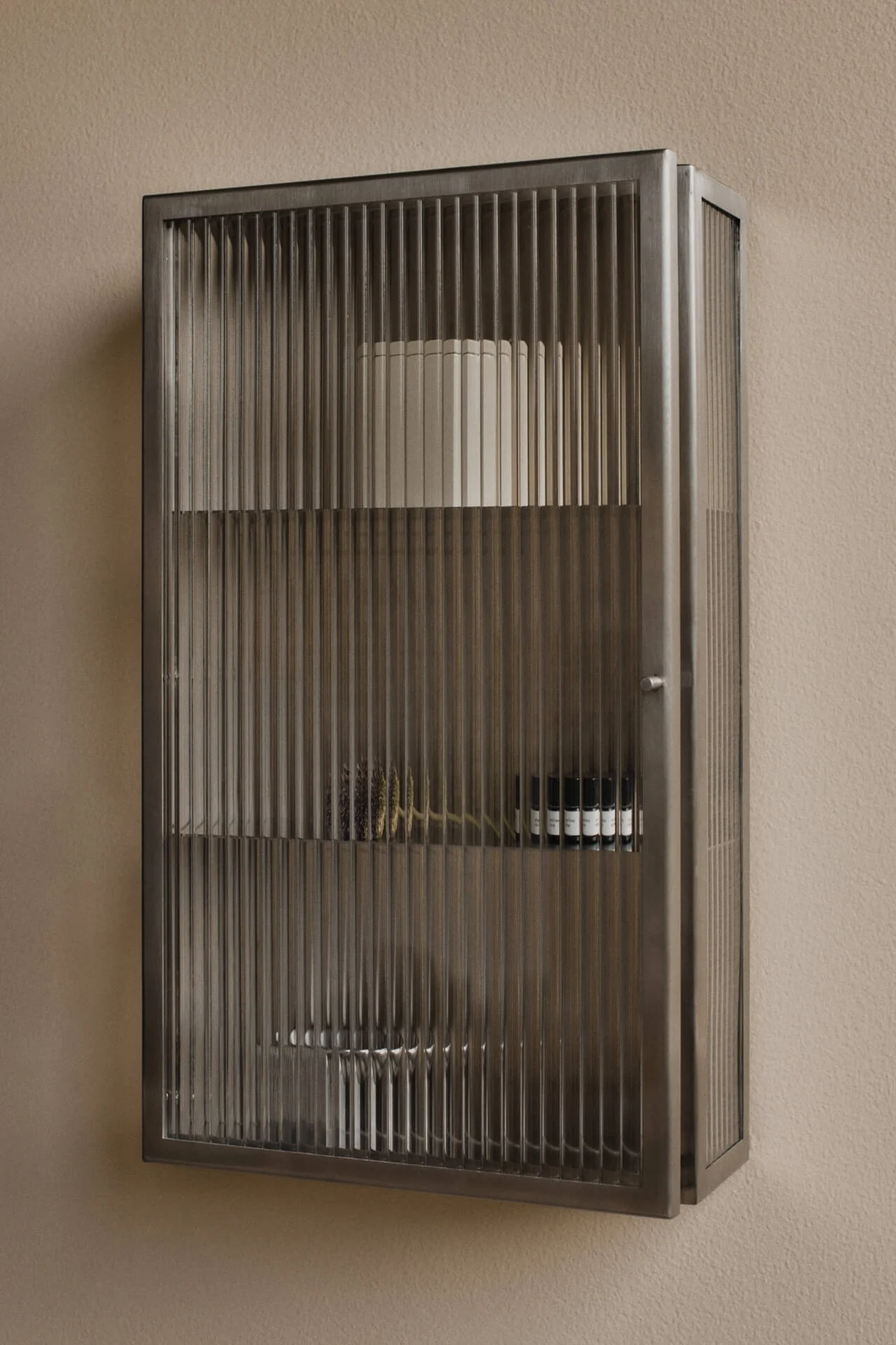 Veggmontert, mørkt metallskap med rillede glassdører på en beige vegg. Inneholder hyller med en hvit sylinder, tørkede botaniske planter og små mørke flasker.