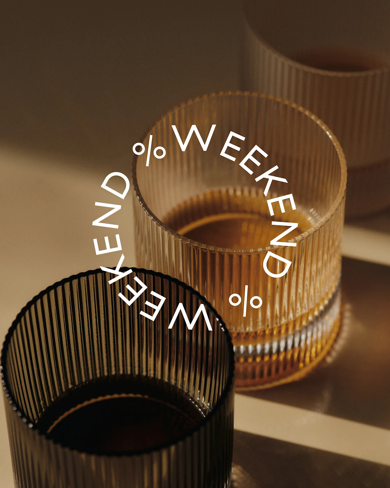 Tre riflede glass, to fylt med mørk væske. Et mørkt glass i forgrunnen, et ravfarget glass bak. Tekst: Weekend %.