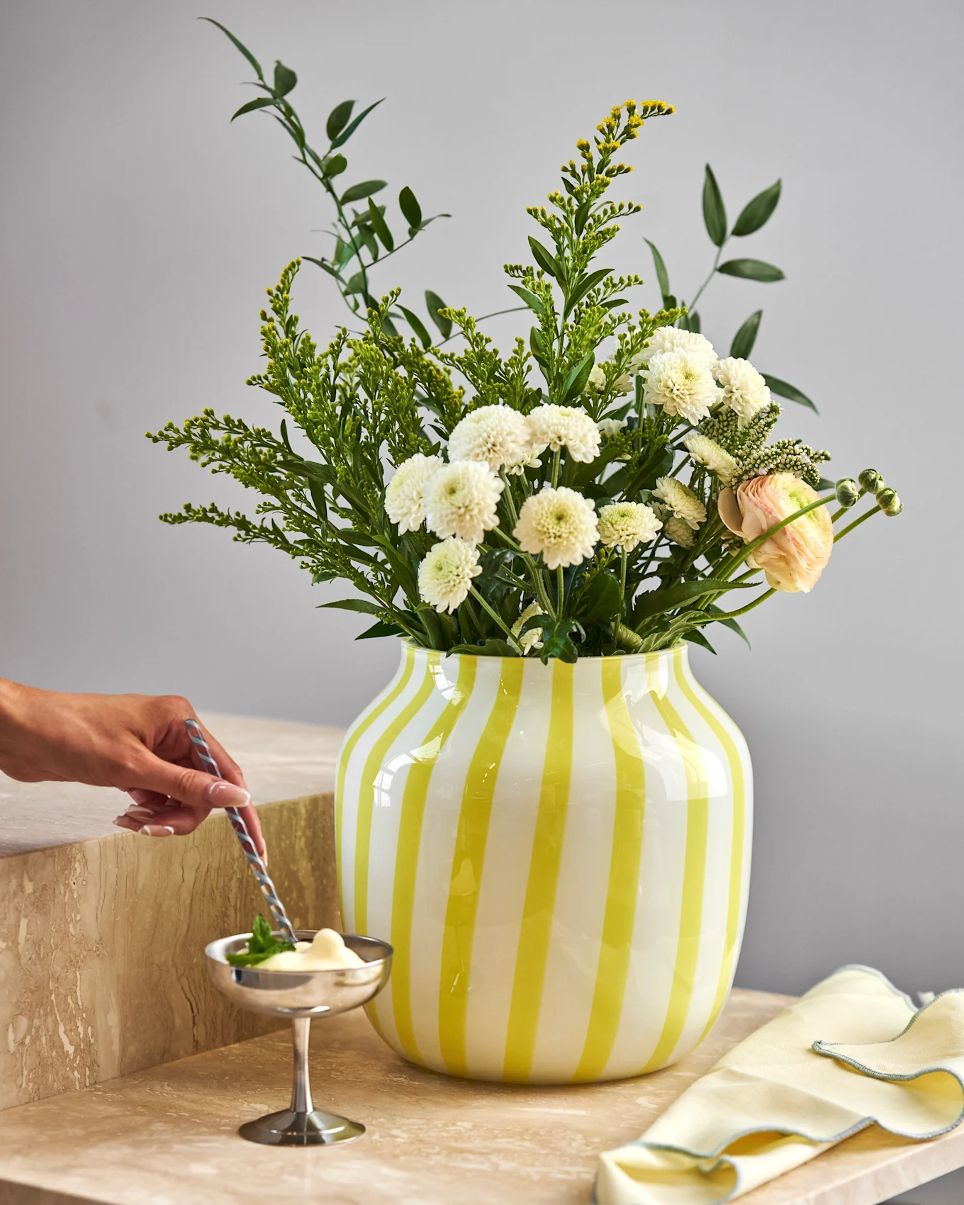 Hånd rører dessert i sølvskål på marmor. Gulstripet vase med hvite krysantemum, gule blomster og rosa ranunkler.