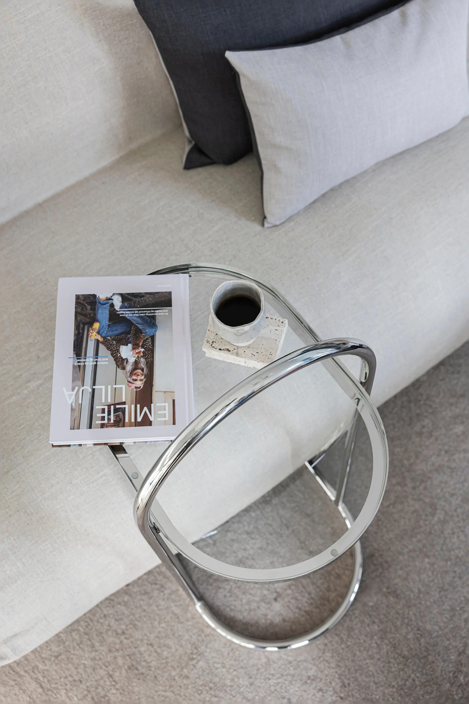 Oversiktsbilde av stue med lys sofa, grå puter og et glass- og krom-sidebord med magasin og kaffe.