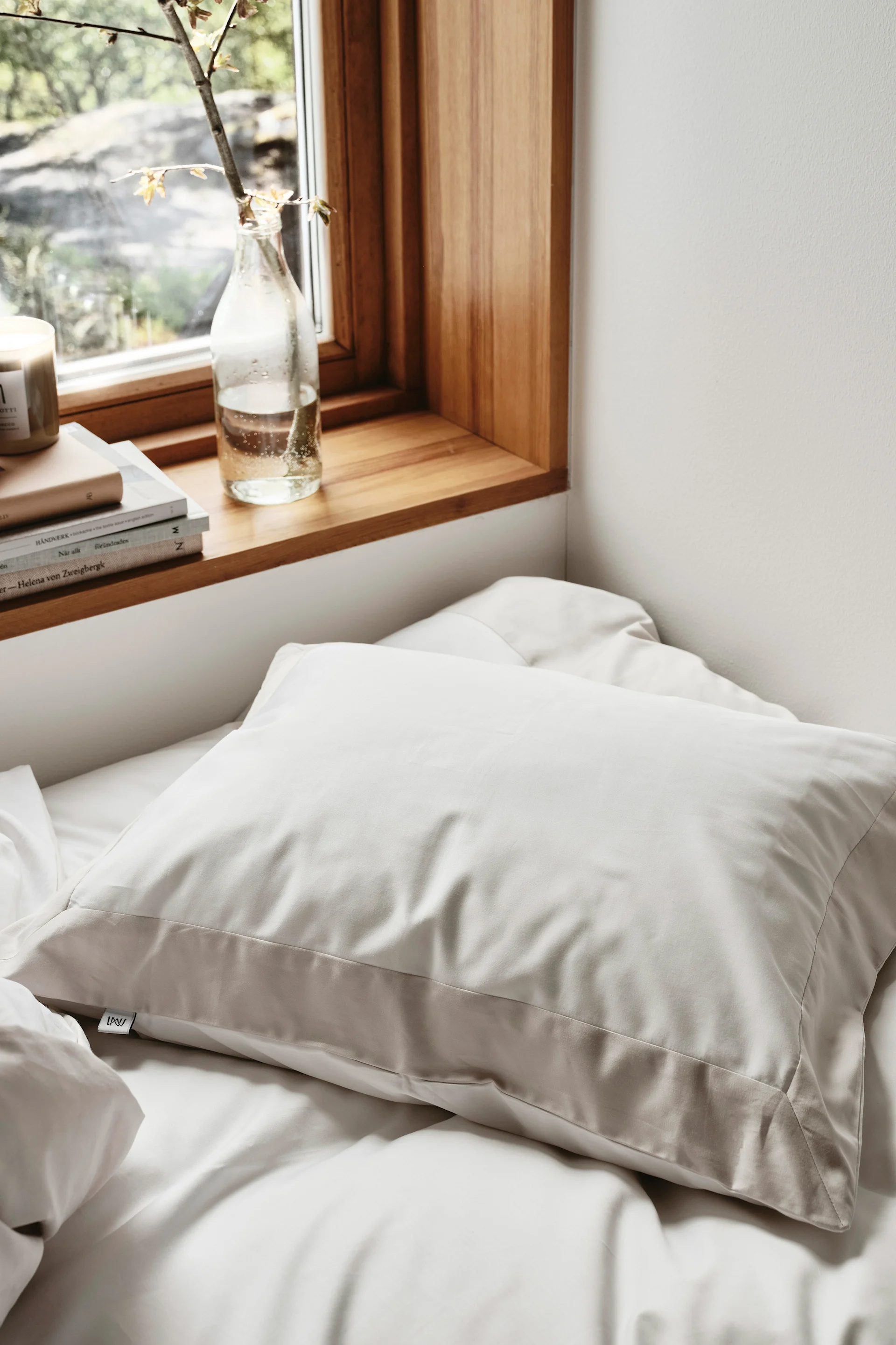 En lys beige pute på en hvit seng, ved siden av et vindu med en trevinduskarm, bøker og en plantegren i en flaske.