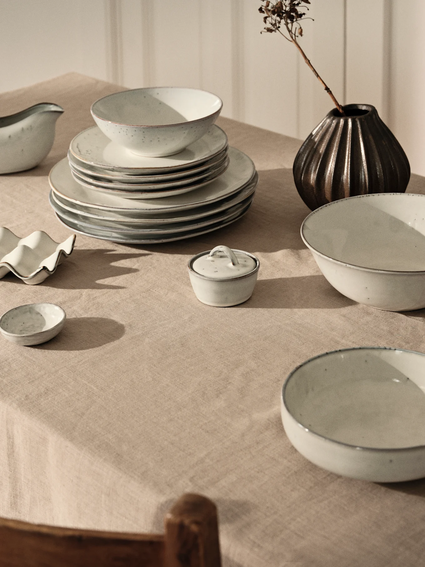 Lys grått keramisk servise med stablede tallerkener og skåler på beige linduk, sammen med en mørk vase med tørket gren.