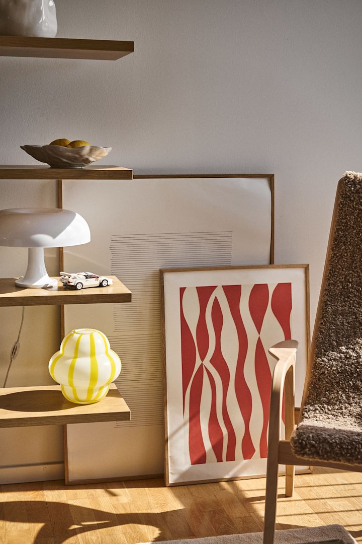 Curlie-vasen fra Byon har et rundt, bulbete design i gult og hvitt stripet glass. Her er den plassert i en åpen bokhylle sammen med Shell-skålen, lamper og malerier.