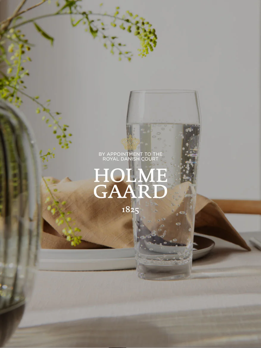 Et høyt glass med kullsyreholdig vann ved siden av en brun serviett og en plante, med Holmegaard-logoen overlagt.