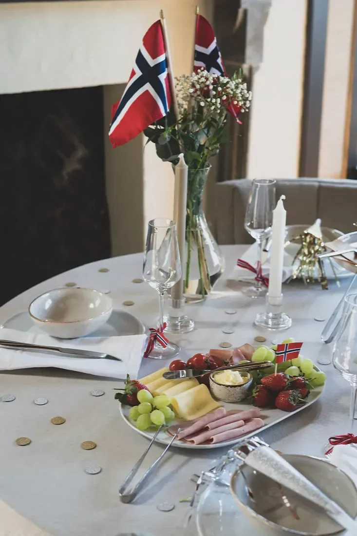 Et festlig dekket bord med norske flagg, blomster, lys og et fat med ost, skinke, frukt og tomater.