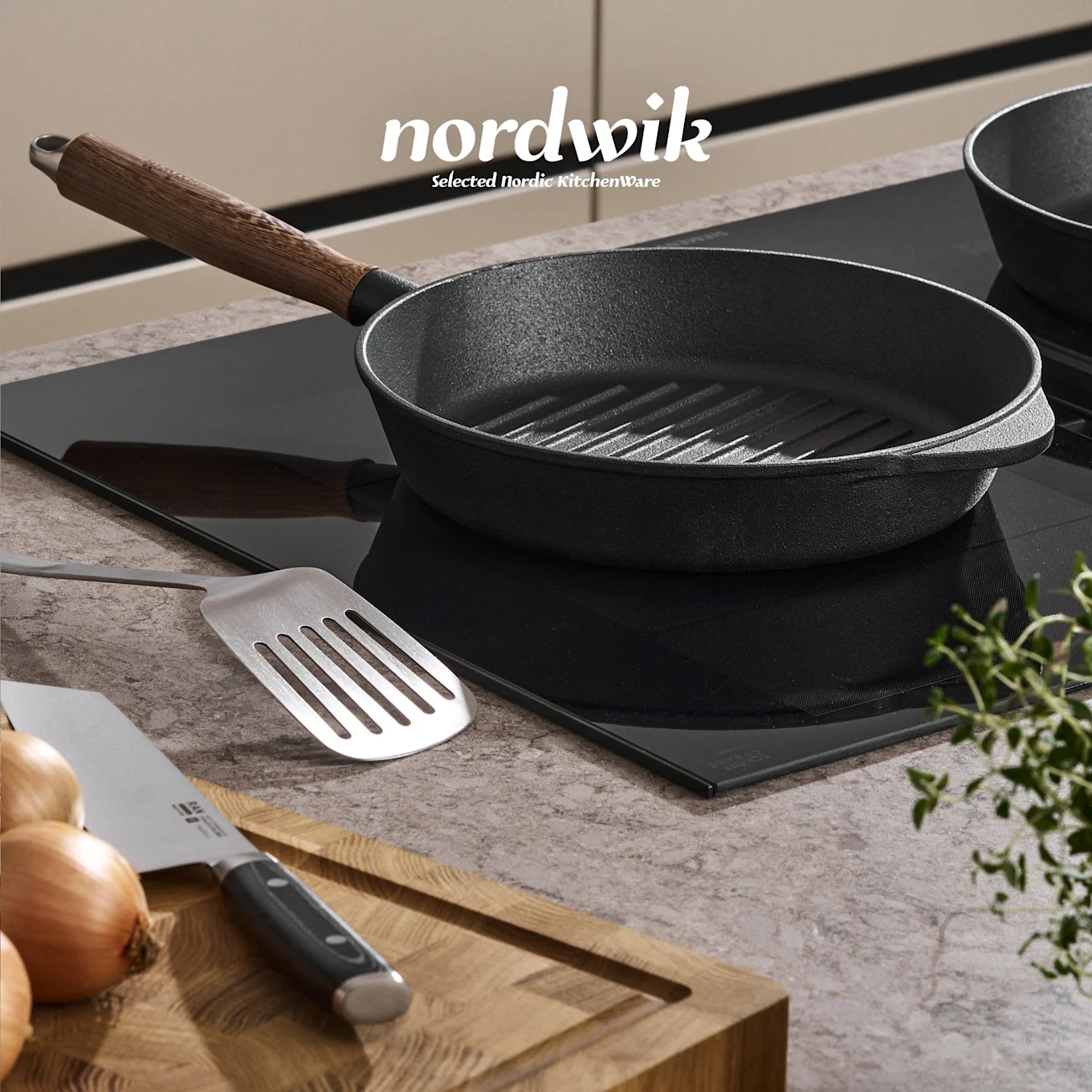 En svart støpejernsgrillpanne på en induksjonstopp, med en stekespade, løk, kniv og timian på et skjærebrett i tre. Nordwik-logo synlig.
