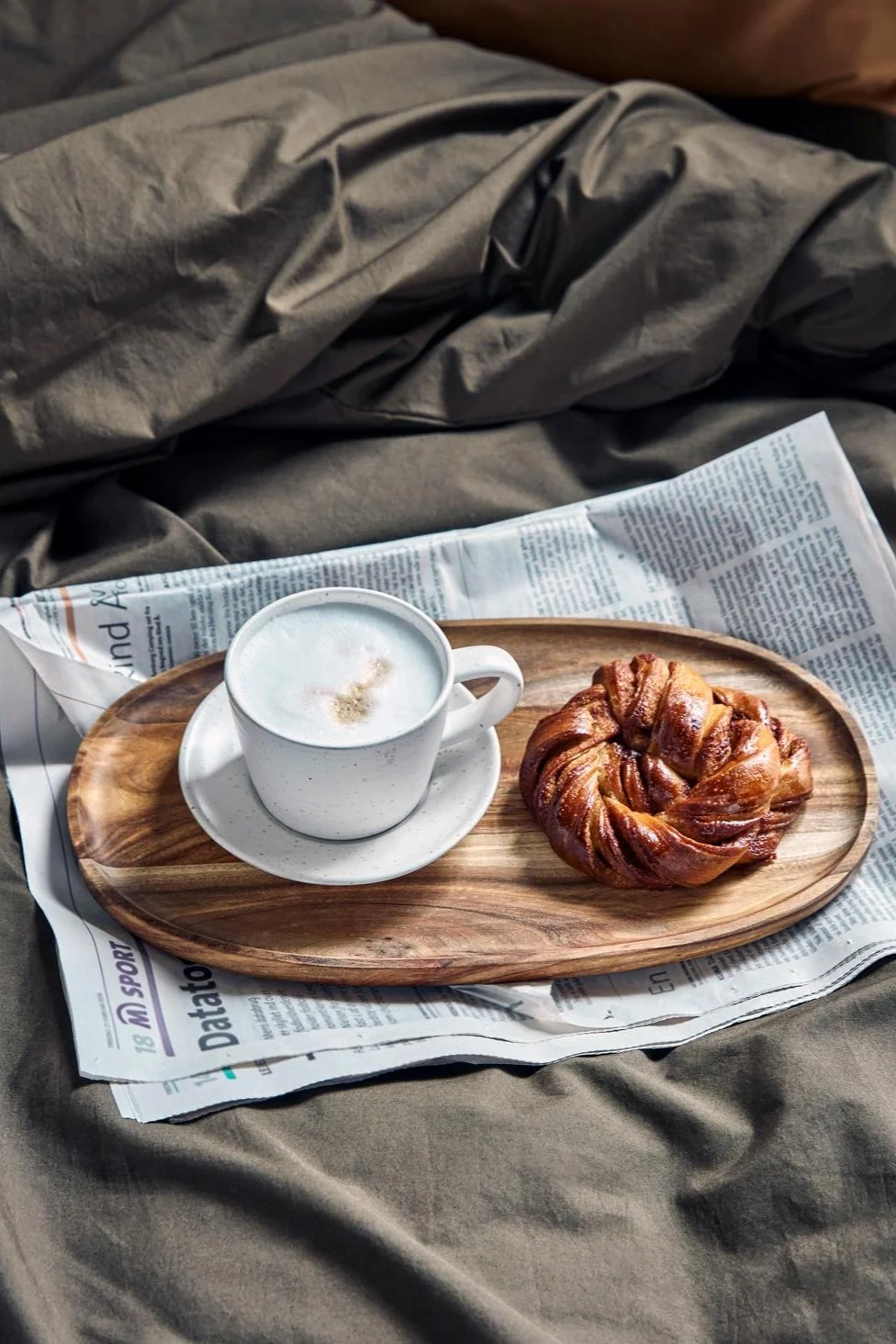 En cappuccino i den hvit-grå Pion-koppen fra House Doctor står på et lite trebrett, oppå en avis, i en seng – ved siden av ligger en svensk kanelbolle.
