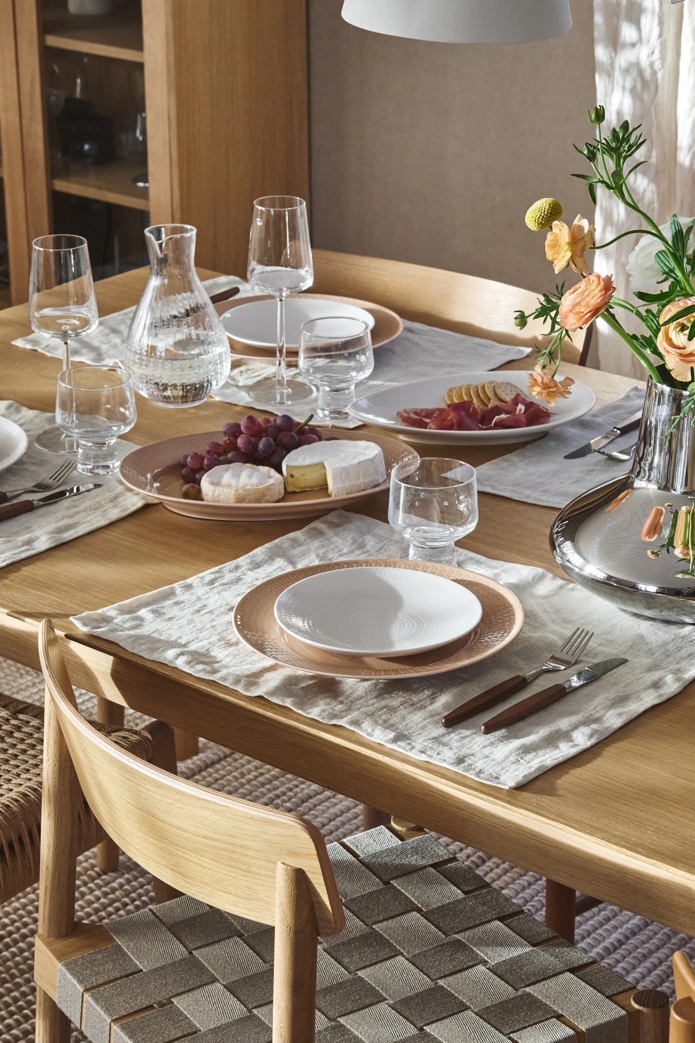 Spisebord i tre med tallerkener, glass, vannkaraffel, druer, oster, spekemat og blomster. Flettede stoler.