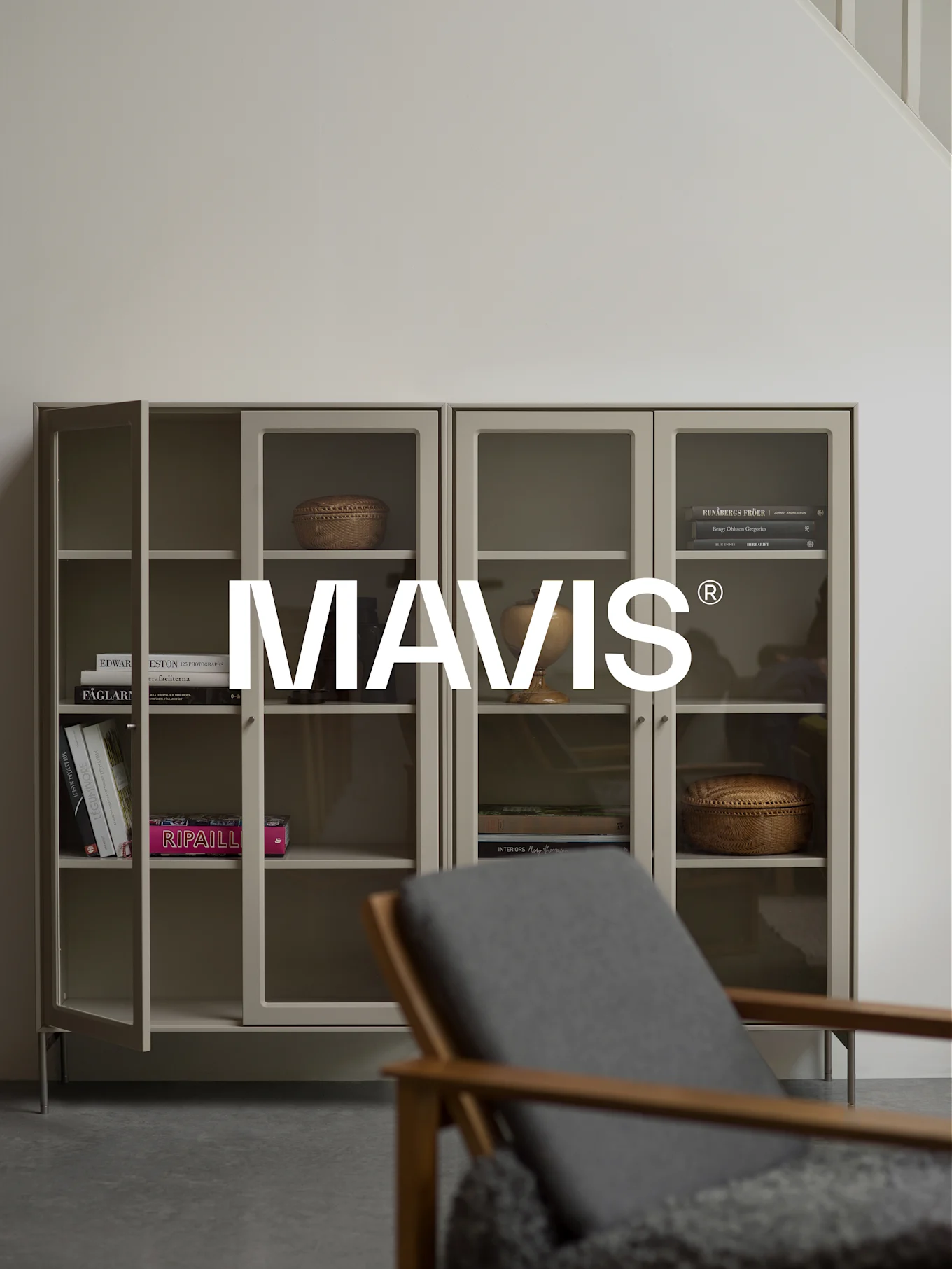 To lysgrå skap med glassdører, som viser bøker og dekor. En trestol er uskarp i forgrunnen. 'MAVIS®'-logoen er sentrert.