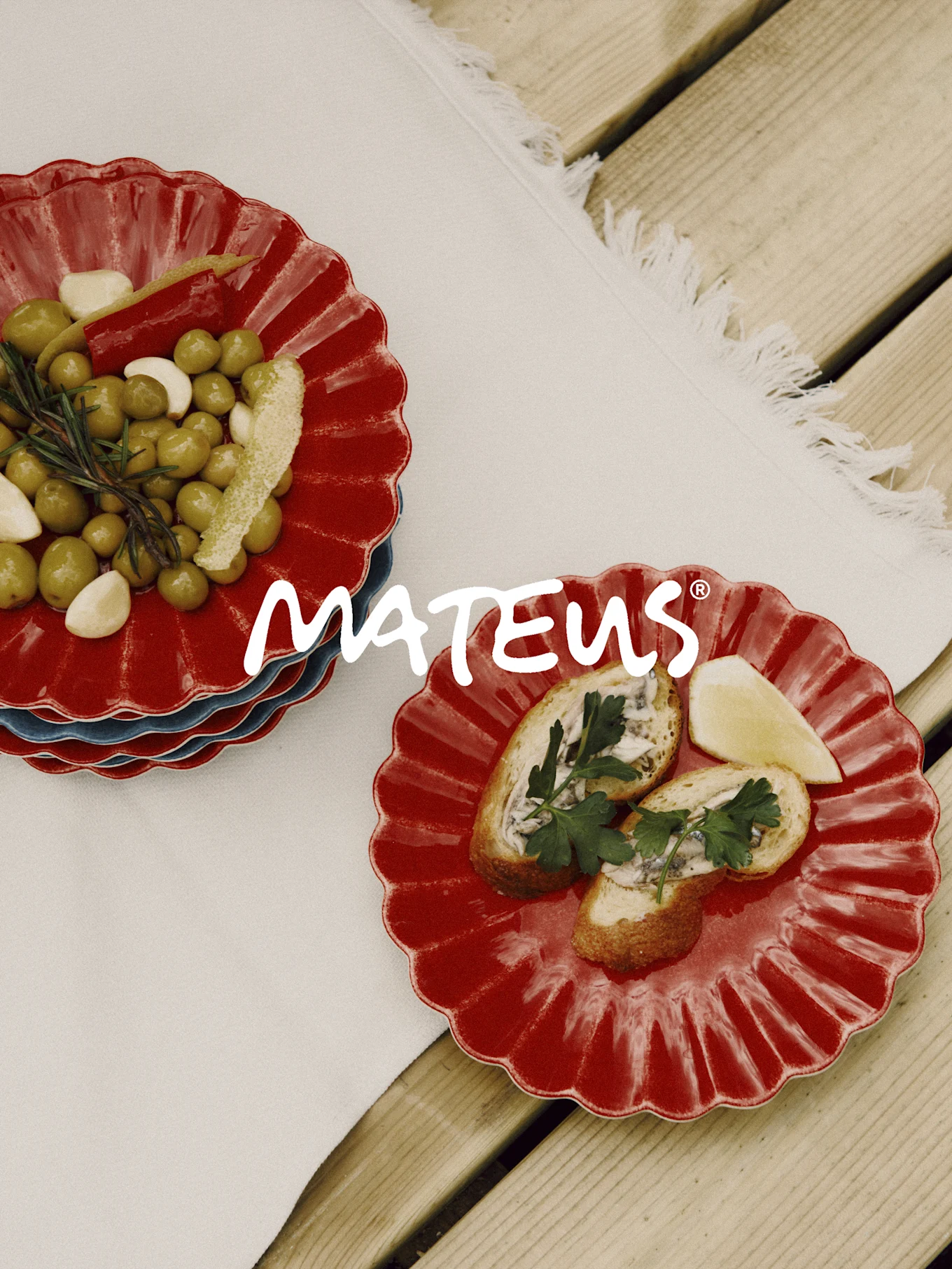 Røde skjellformede tallerkener med oliven, hvitløk, rosmarin og brød med ansjoser på et trebord. MATEUS-merke.