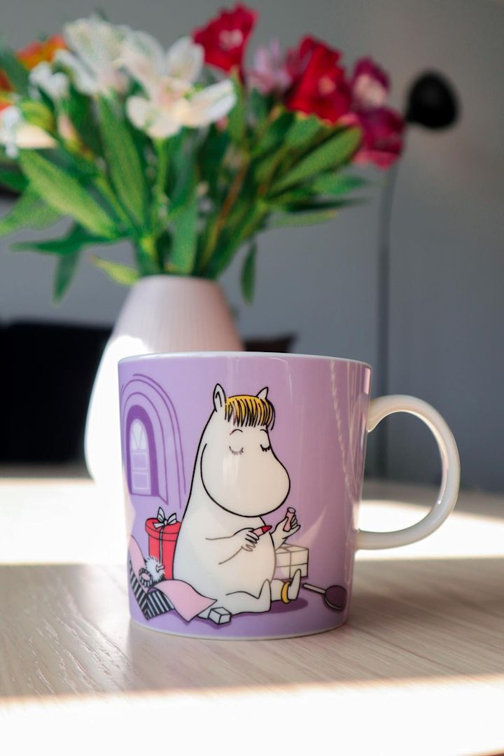 Den lilla mummikoppen med Snorkfrøken på står på et bord, og i bakgrunnen står en vase med en bukett blomster.
