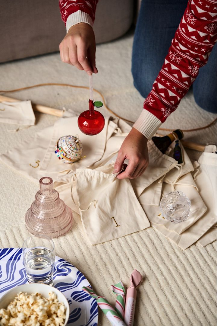 Hendene legger en rød epledekorasjon fra Iittala i posen til en adventskalender fra ferm LIVING.