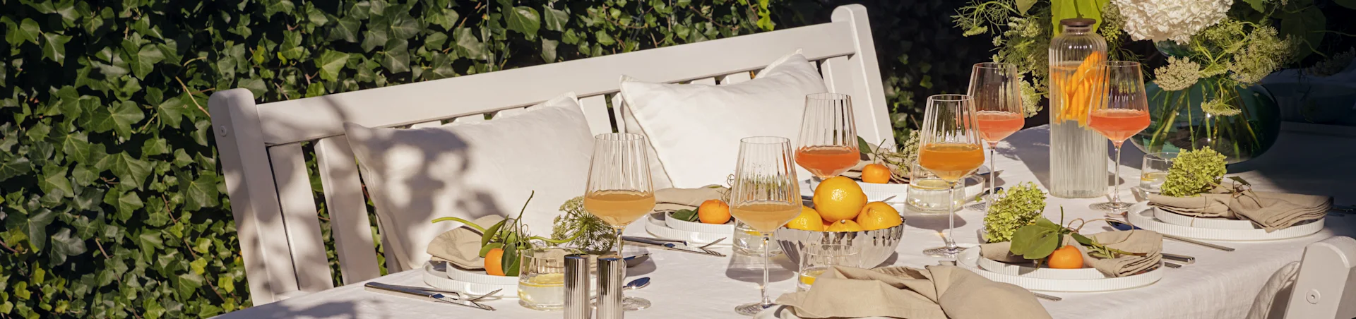 Utendørs bord dekket til sommermåltid med hvite rillede tallerkener, linservietter, riflede glass med drikke, sitroner og mandariner.