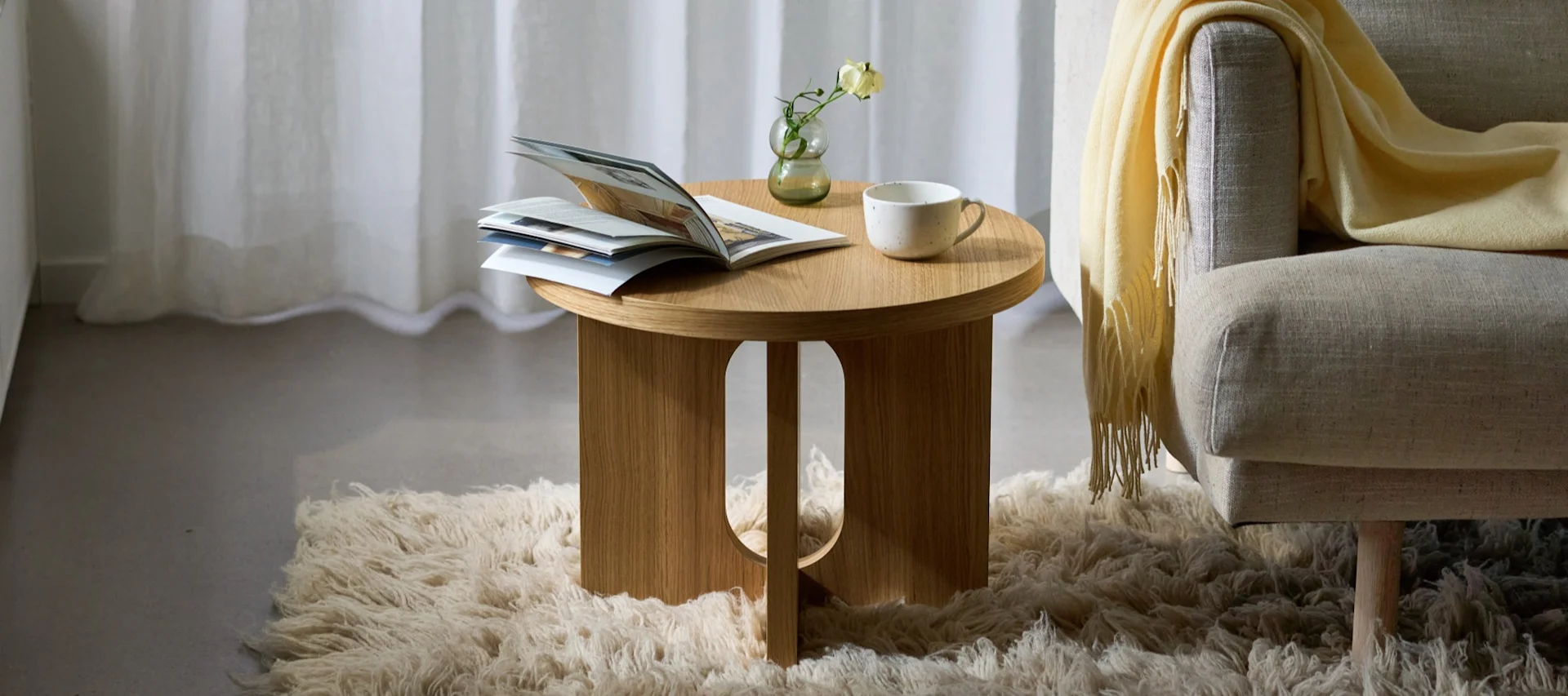 Rundt sidebord i tre med magasin, vase og kopp, ved siden av en lenestol med gult teppe på et lodden teppe.