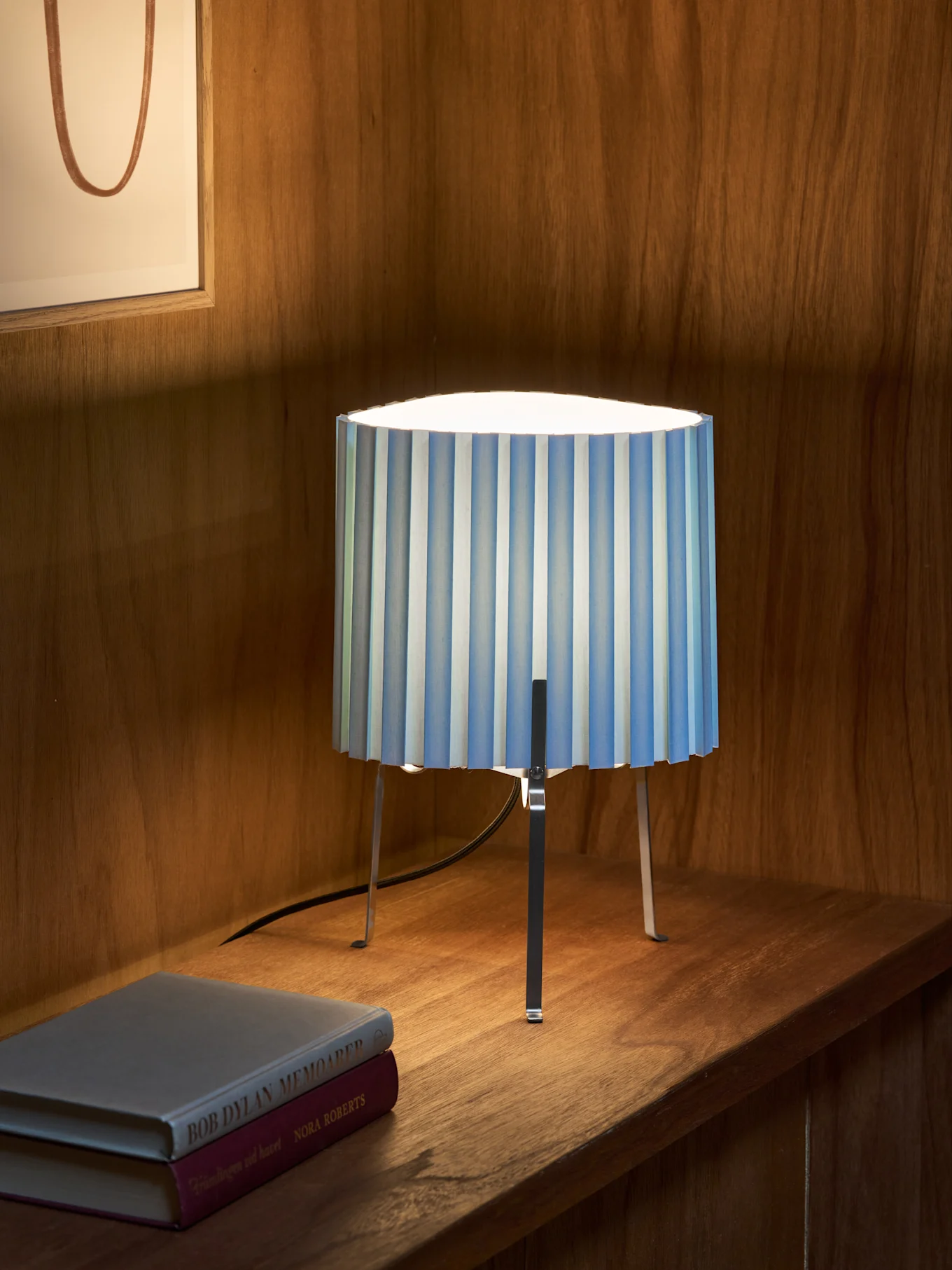 En blå- og hvitstripet plissert bordlampe med tre metallben står på en trehylle ved siden av to bøker. Innrammet kunst i bakgrunnen.