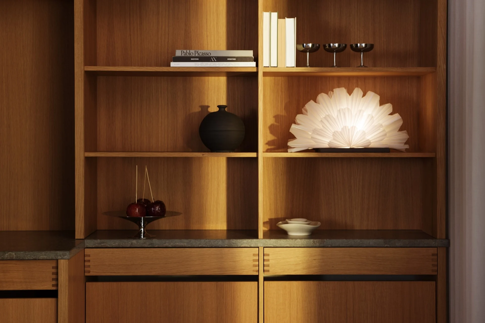 Et moderne treskap med hyller som viser bøker, en svart vase, tre sølvglass, en glødende vifteformet lampe, karamellepler og hvite keramiske fat.