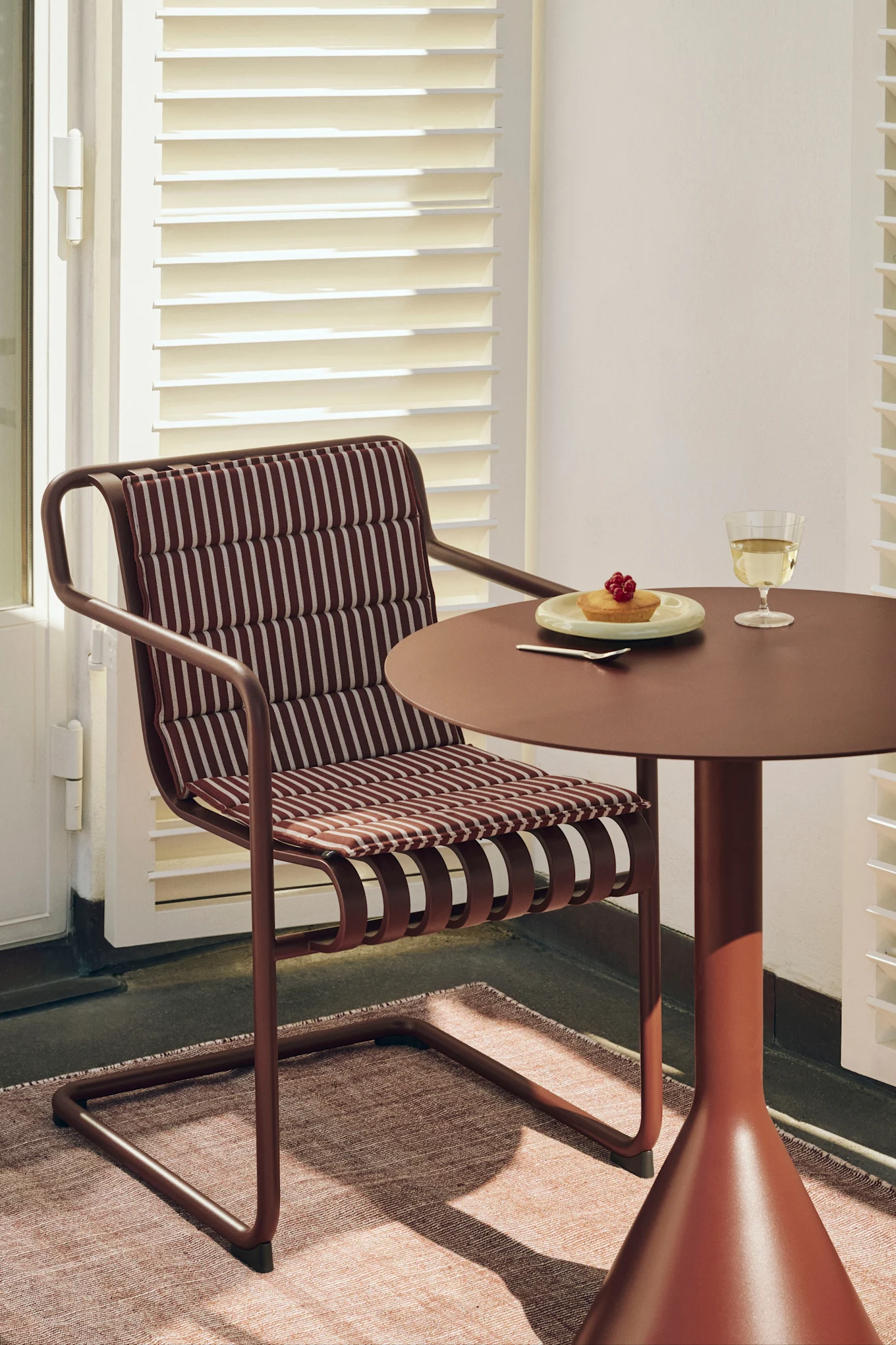 En rødbrun stol med stripete pute ved et matchende rundt bord med en kake med bær og et glass hvitvin.