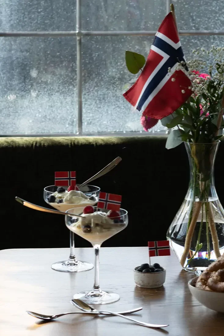 To dessertglass med is, bær og små norske flagg, sammen med en vase med blomster og et stort norsk flagg.
