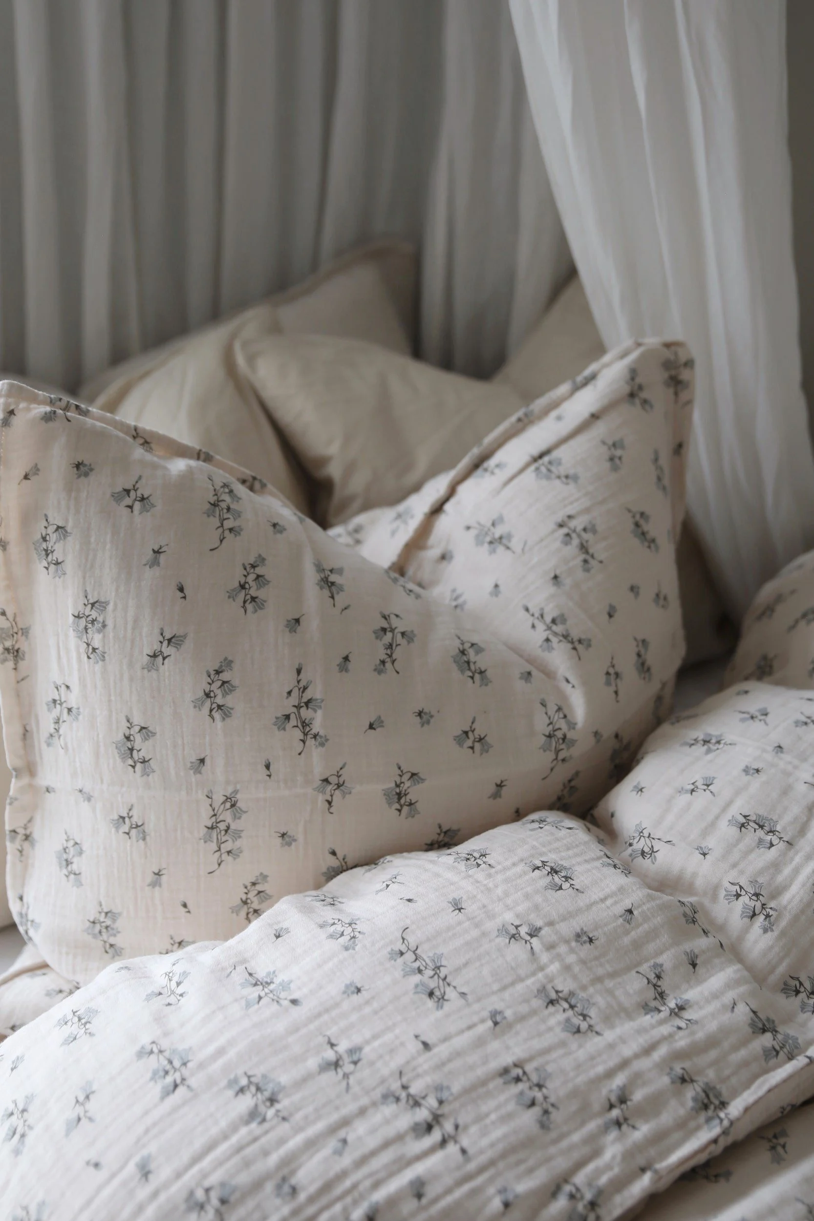 Koselig sengetøy i lyst beige kreppstoff med et delikat blått blomstermønster og hvite gardiner.