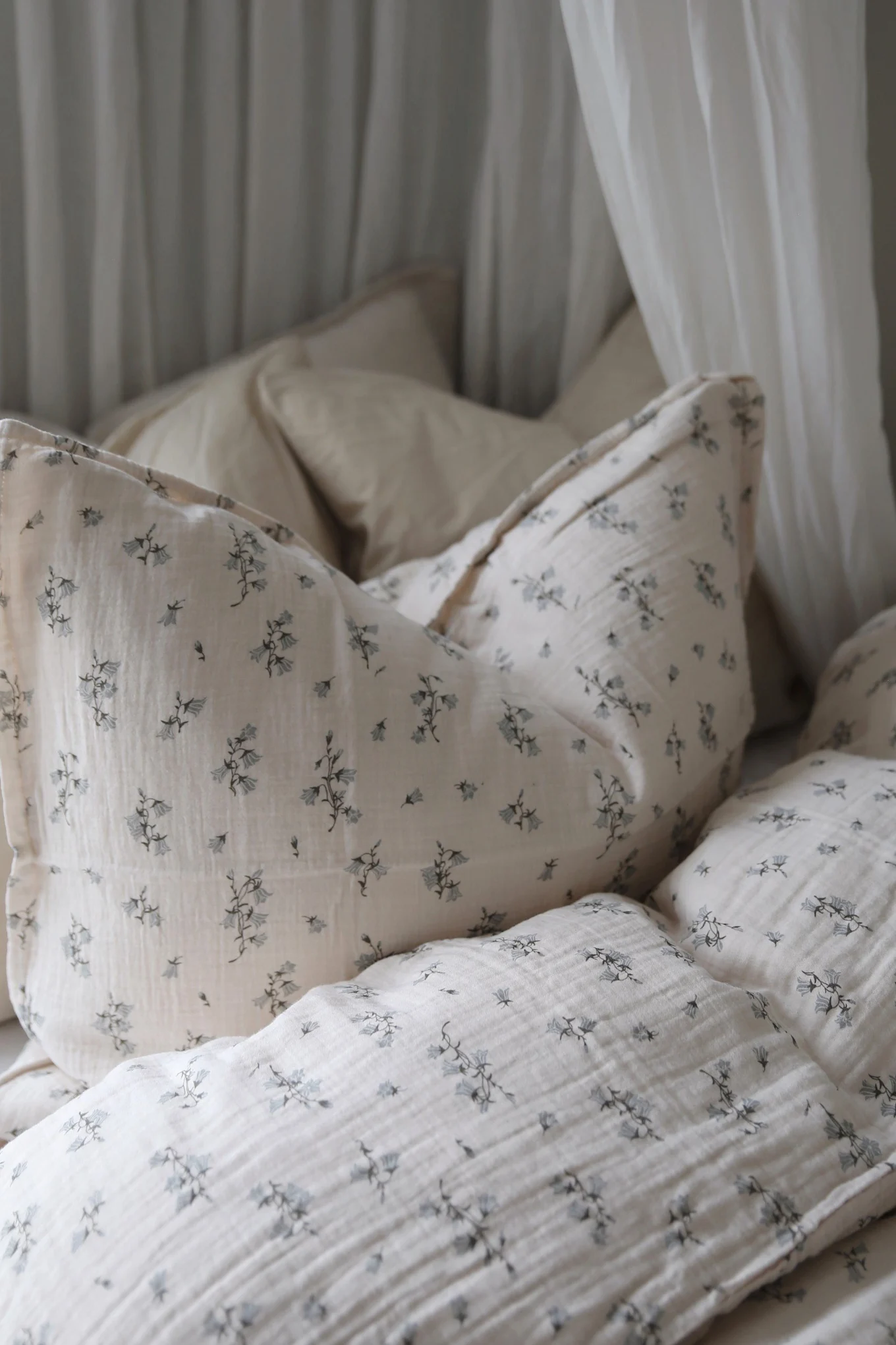 Koselig sengetøy i lyst beige kreppstoff med et delikat blått blomstermønster og hvite gardiner.