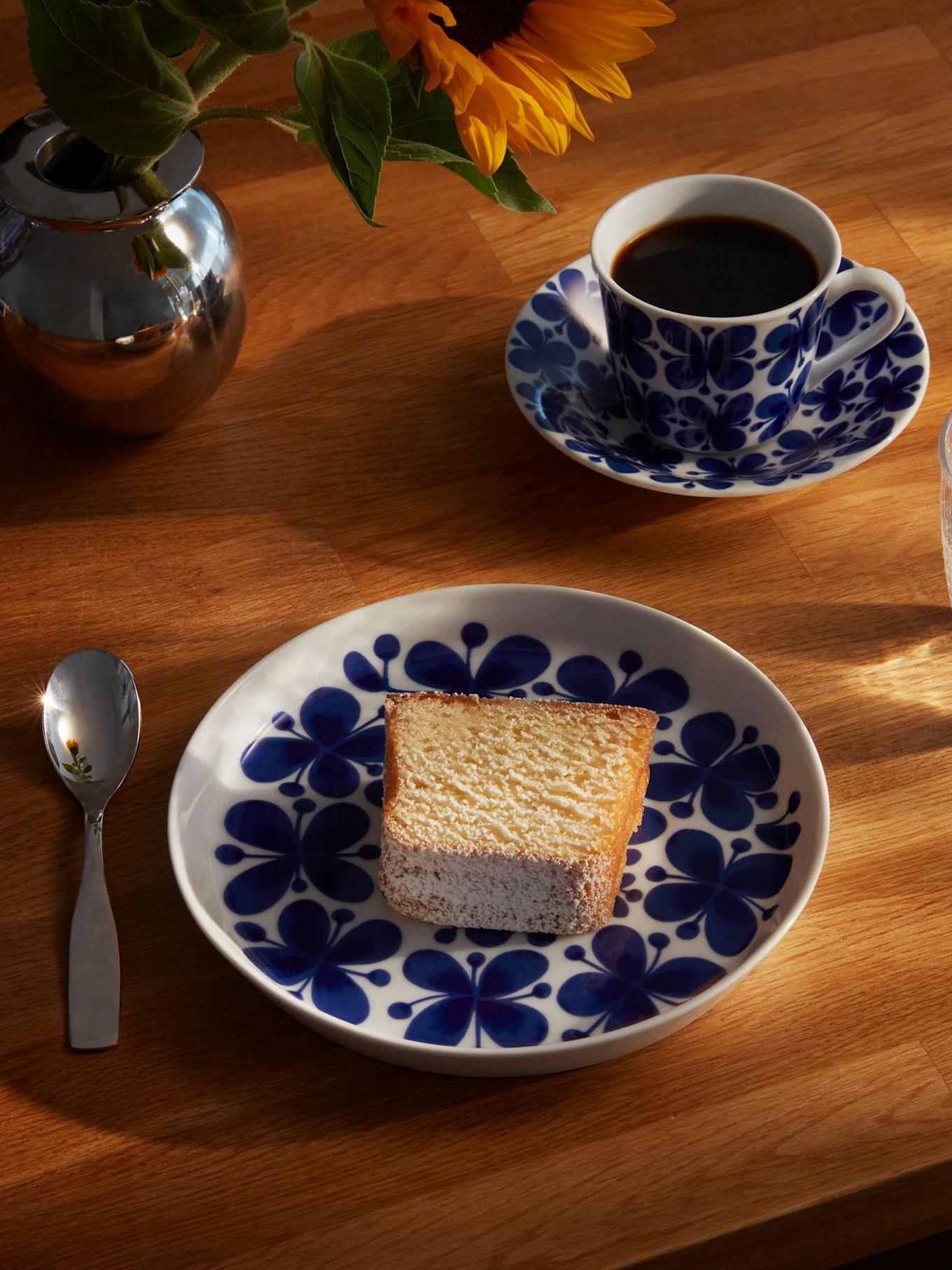 Et stykke kake på en blå og hvit blomstret tallerken, med en kaffekopp, et glass vann, en skje og en solsikke i en sølvvase på et solfylt trebord.